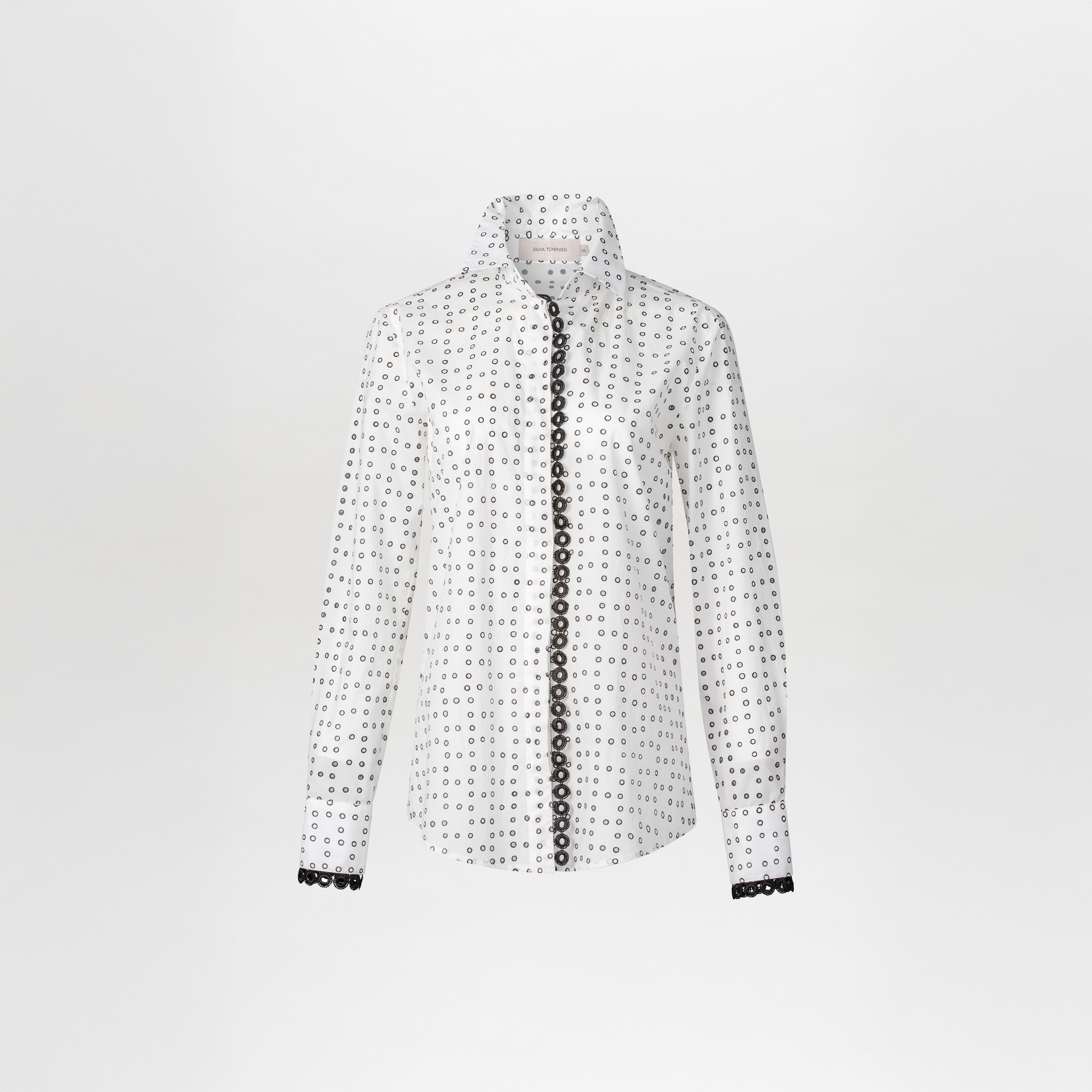 A woman stands against a blue background, wearing the white Naroa Blouse with black dots and light gray pants, accessorized with a chain belt, ready for the fresh styles of September 2nd, 2024.