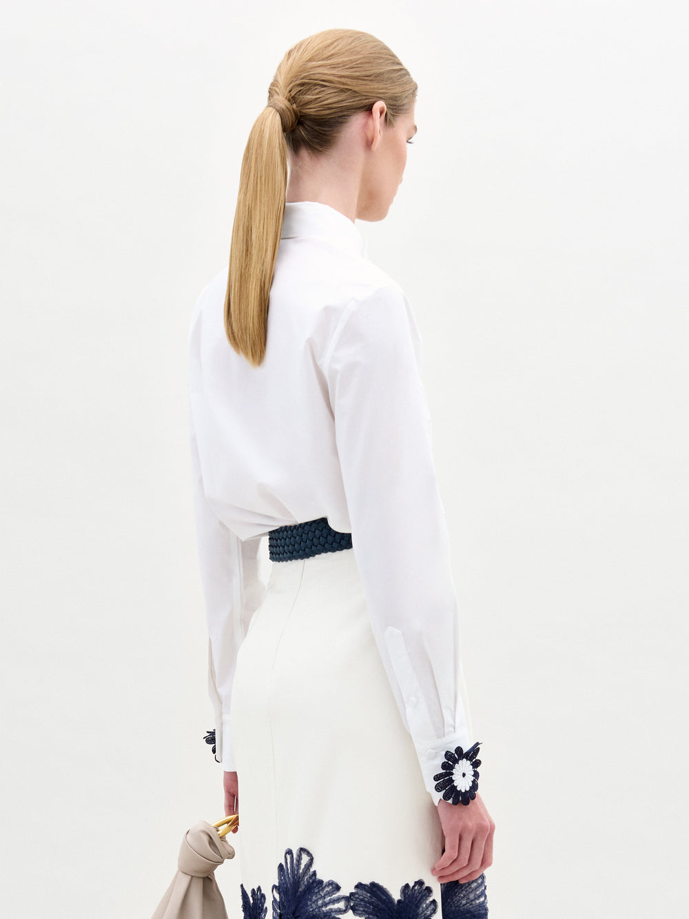 A woman wears the Naroa Blouse—a white top with floral appliqué details—paired with a white Italian-fabric skirt, standing against a plain light background.