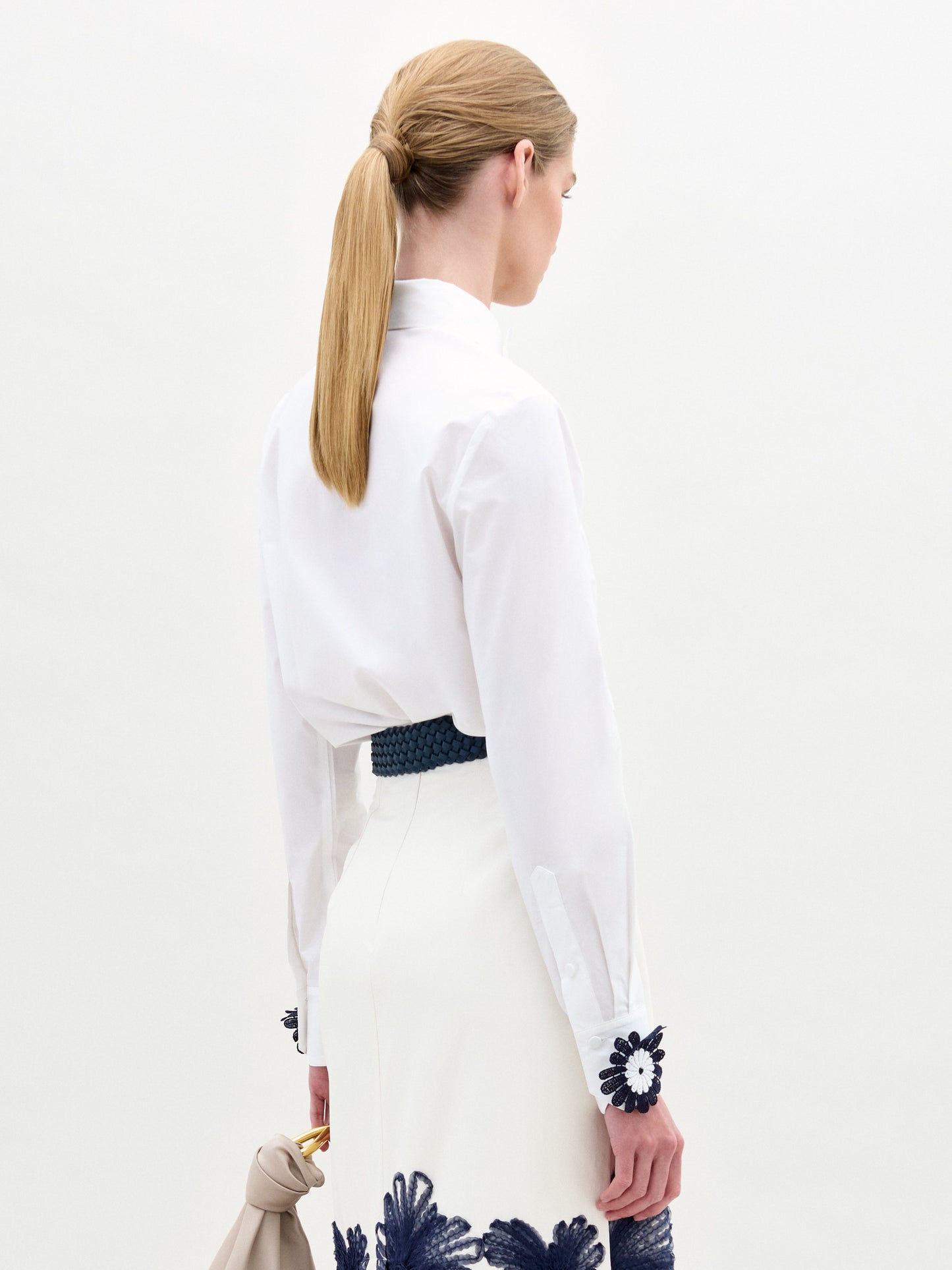 A woman wears the Naroa Blouse—a white top with floral appliqué details—paired with a white Italian-fabric skirt, standing against a plain light background.