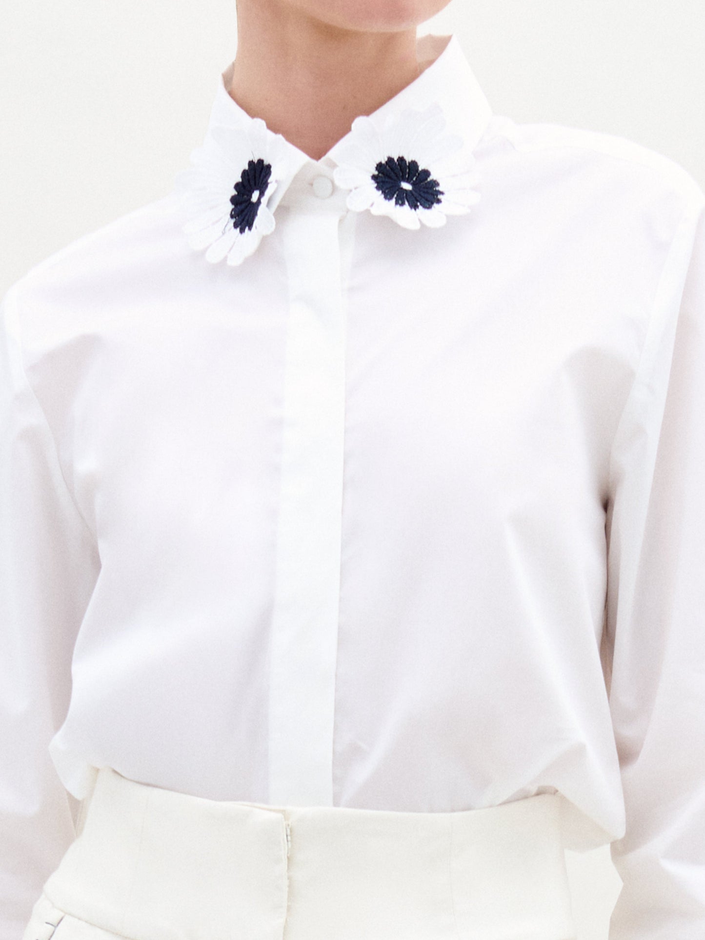A woman wears the Naroa Blouse—a white top with floral appliqué details—paired with a white Italian-fabric skirt, standing against a plain light background.