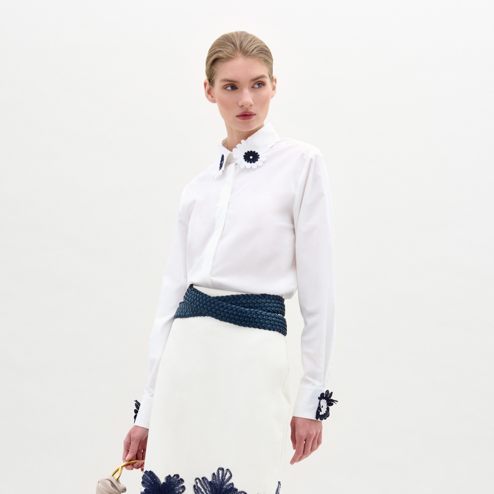 Woman wears the Naroa Blouse, a white long-sleeve shirt with dark floral appliqué accents, paired with a matching skirt, navy braided belt, and light handbag against a plain background.