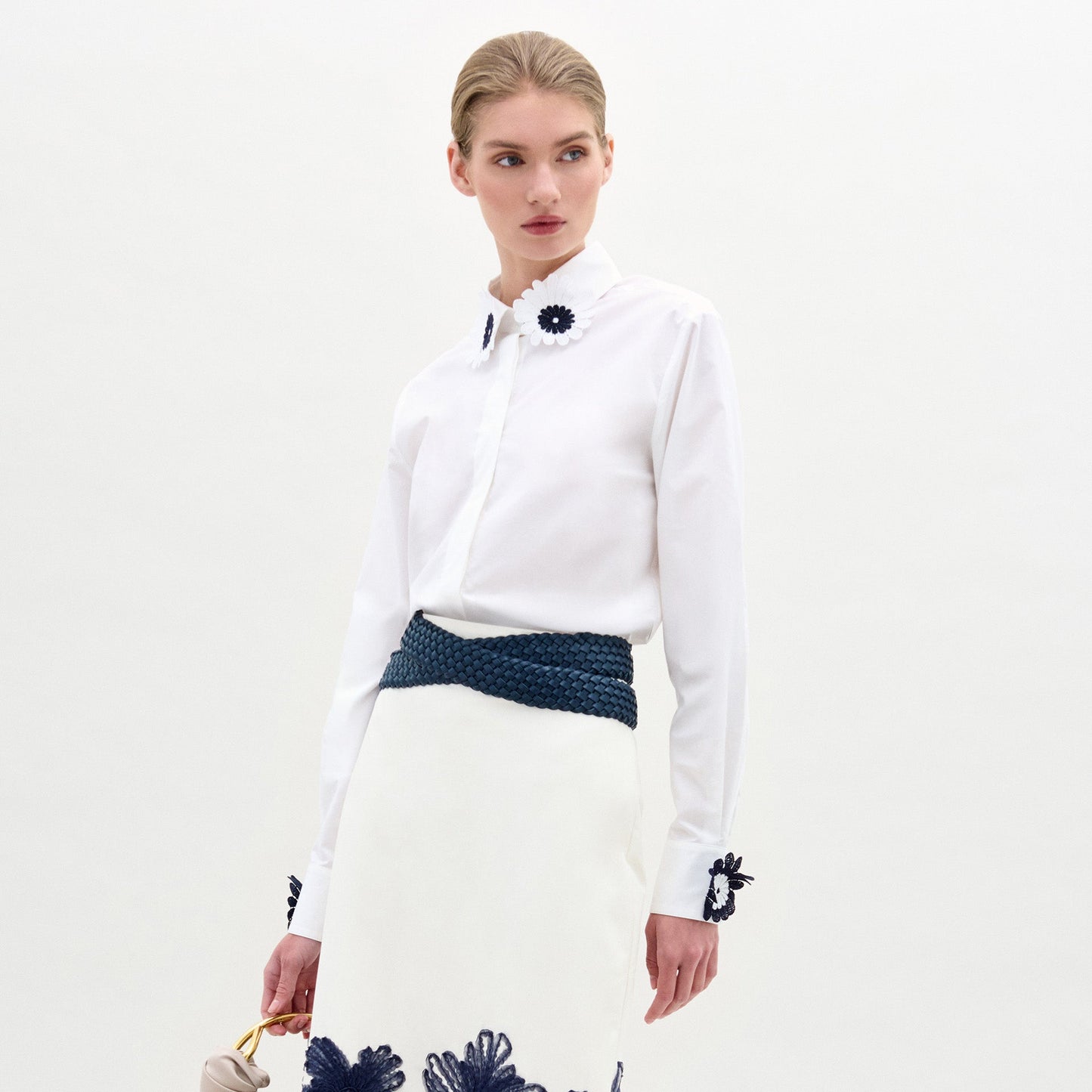 A woman wears the Naroa Blouse—a white top with floral appliqué details—paired with a white Italian-fabric skirt, standing against a plain light background.