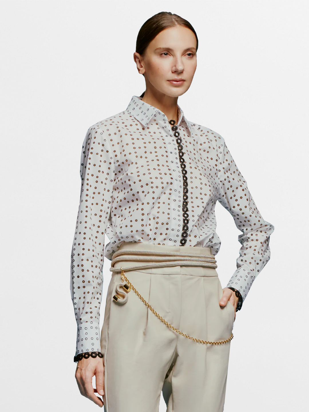 A woman stands against a blue background, wearing the white Naroa Blouse with black dots and light gray pants, accessorized with a chain belt, ready for the fresh styles of September 2nd, 2024.