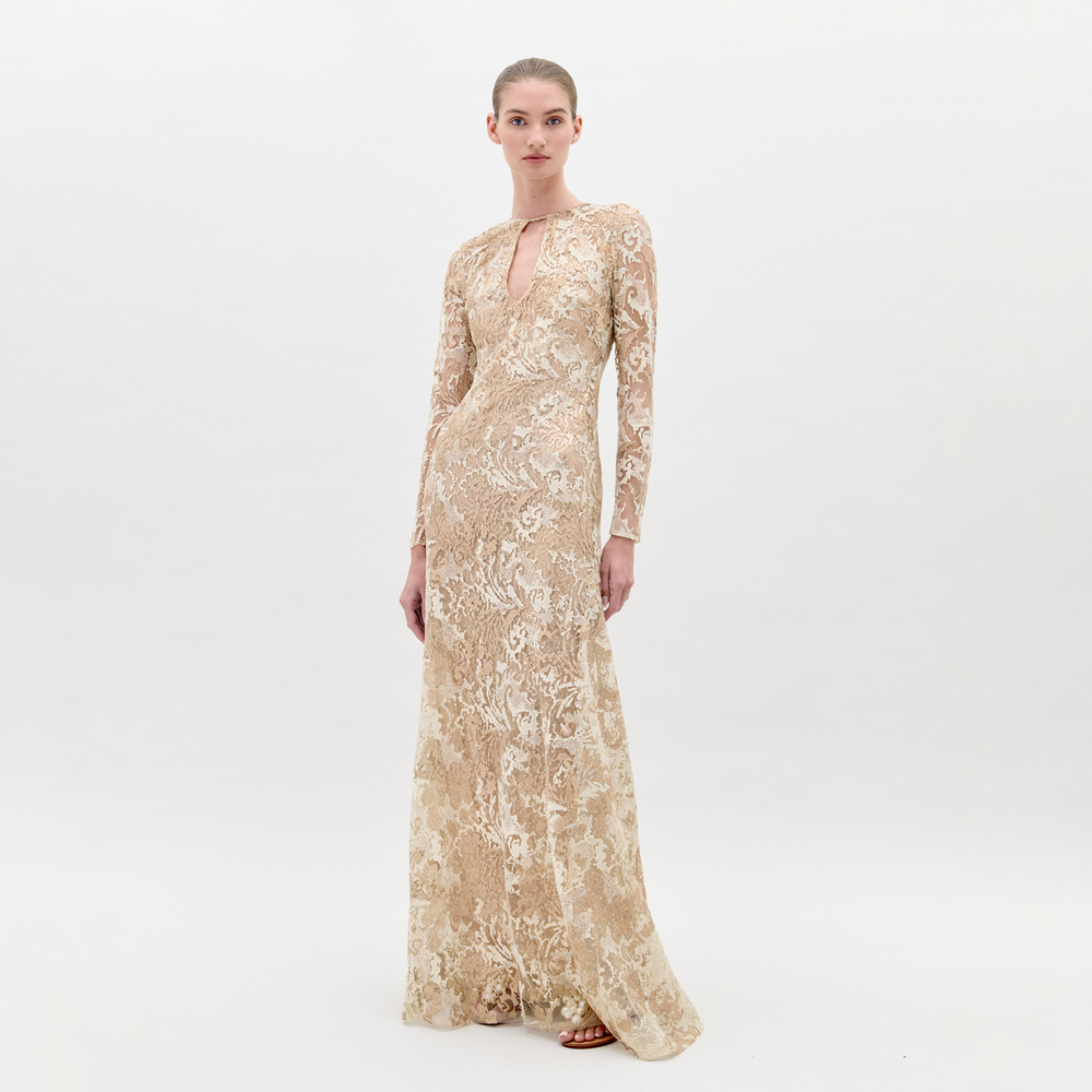 A woman in the Nila Dress Gold, a beige, floor-length, long-sleeved lace gown with a keyhole neckline, stands against a plain background, ready for pre-order ahead of the ship-by date on February 1st, 2025.
