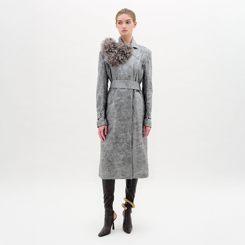 A woman stands against a plain background wearing the Nilo Trench, a textured gray structured coat with a large floral shoulder detail, paired with black heeled boots and a gold ankle accessory.
