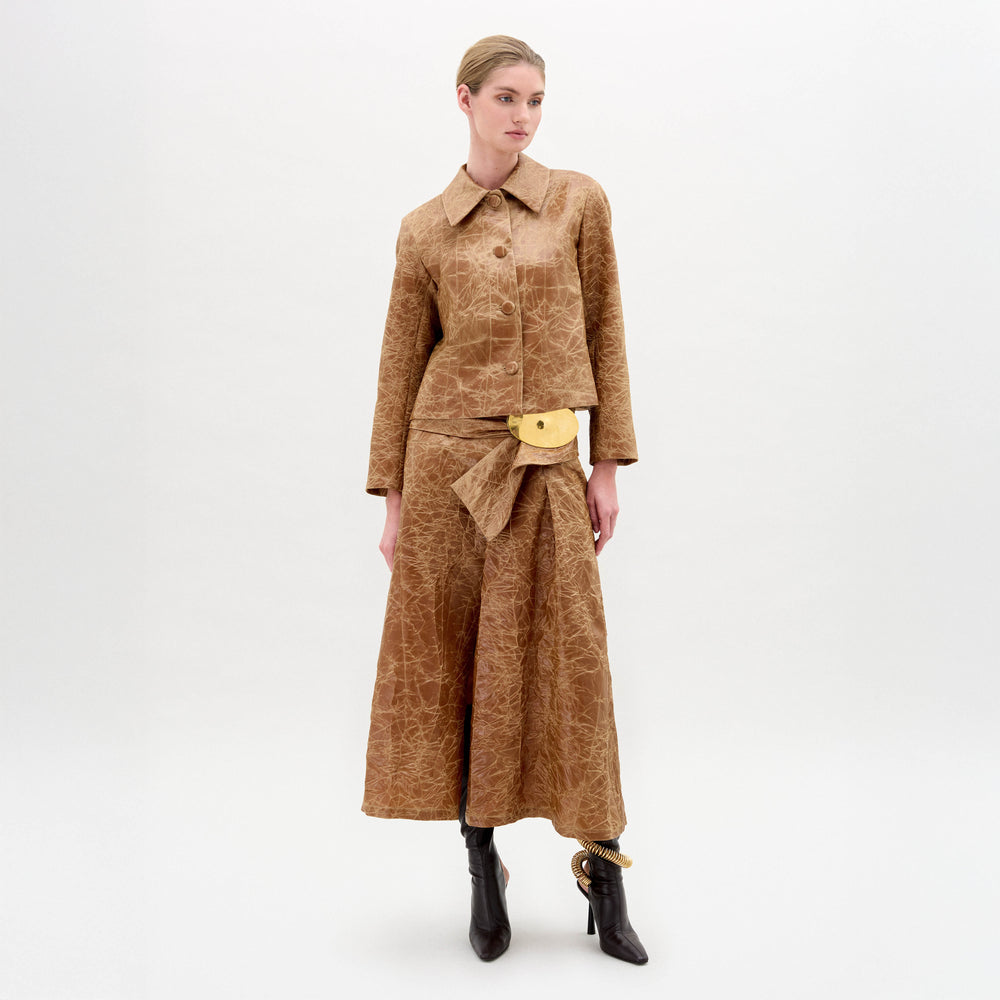A woman wears the Olga Skirt in brown with a matching textured A-line jacket, gold accent belt, dark ankle boots, and a minimal background.