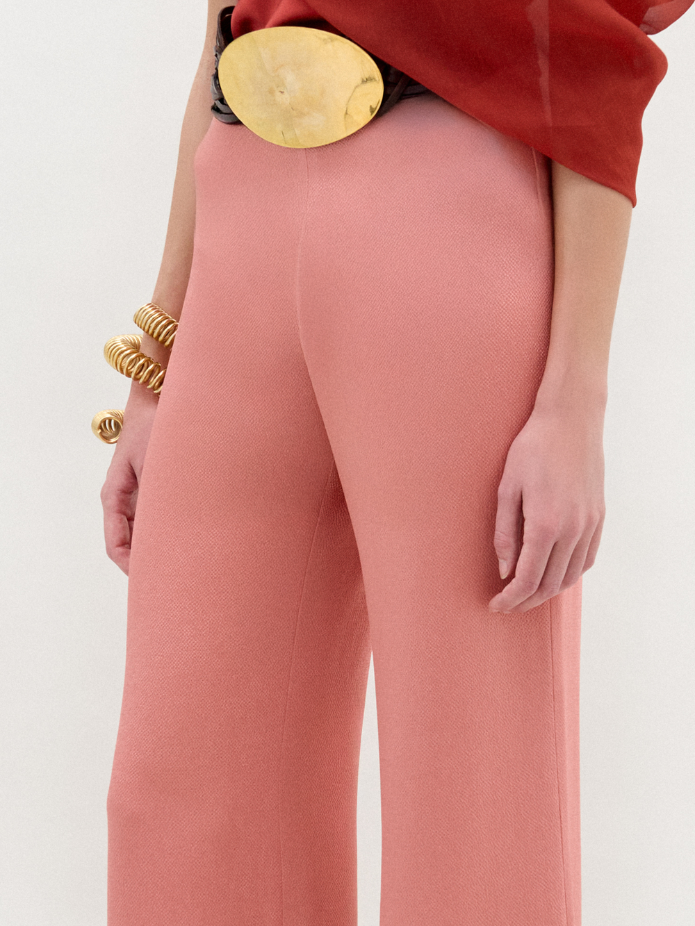 A woman stands in high-waisted pink Palermo Pants, crafted from luxurious Italian fabric, paired with a red one-shoulder top, gold belt, gold bracelets, and open-toe shoes against a white background.