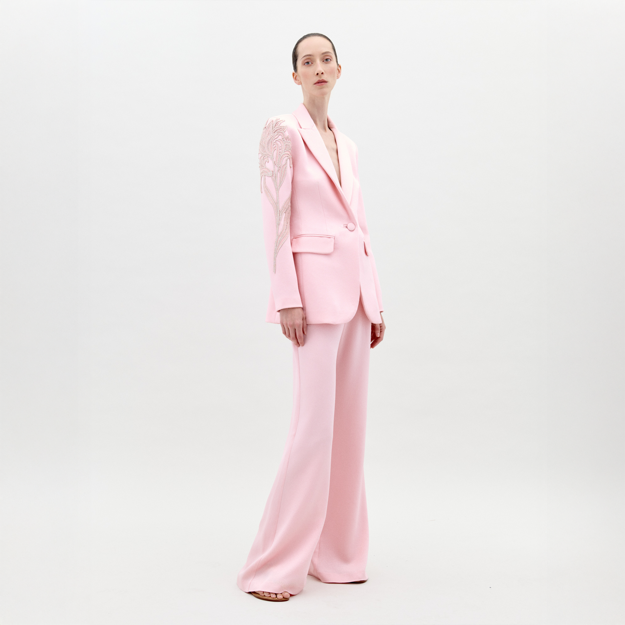 A person stands wearing the Palermo Pant Pink pantsuit, featuring floral detailing on the left shoulder and sleeve, against a plain white background. Available for pre-order now, this elegant outfit will have an exclusive ship-by date of February 1st, 2025.