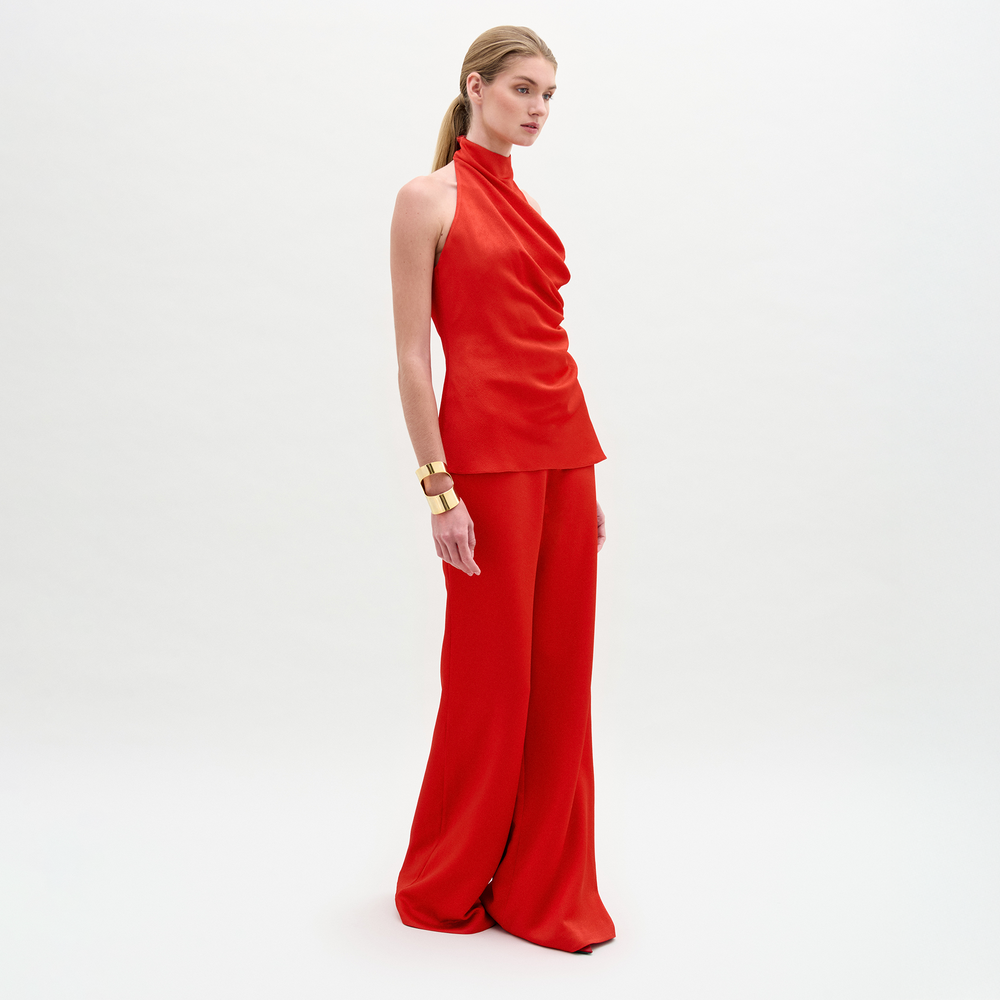 A woman poses against a white background in the sleeveless red halter top and high-waisted, wide-leg Palermo Pant in hammered satin, accessorized with gold bracelets.