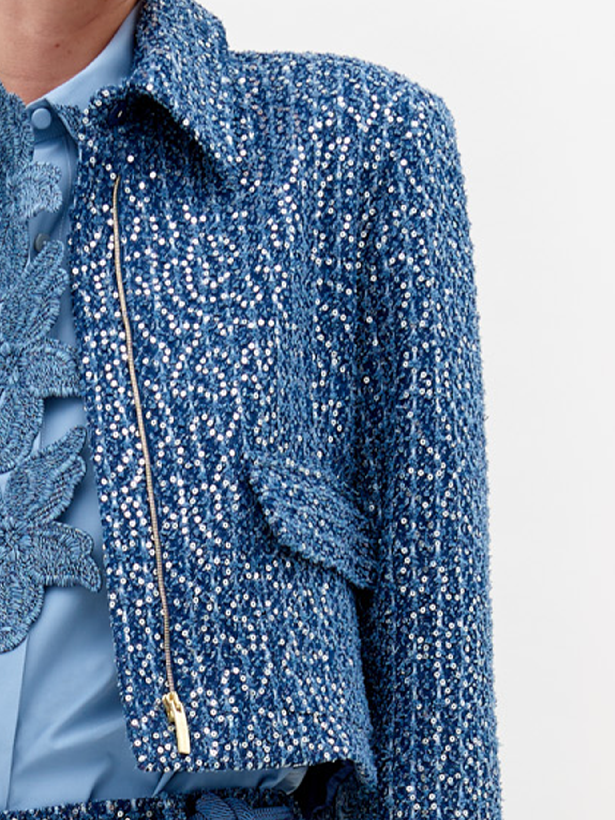 A person wearing a Perla Jacket Navy Sequins and pants, with a light blue shirt and a belt with pearl-like decorations, stands against a plain white background. Pre-order now to ensure delivery by November 15th, 2024.