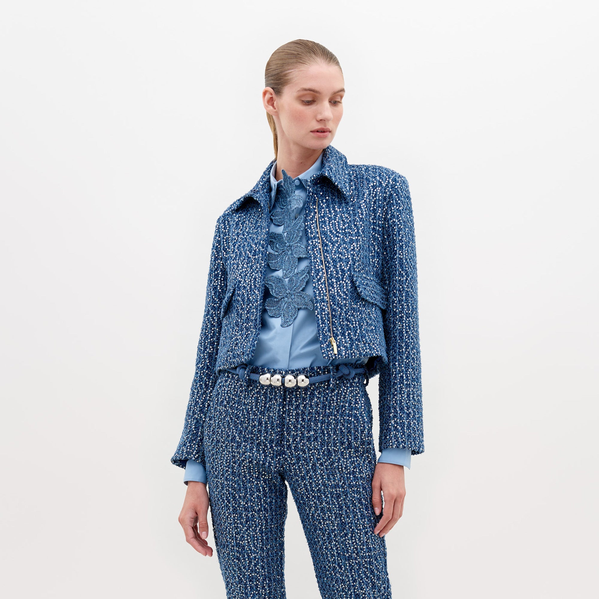 A person wearing a Perla Jacket Navy Sequins and pants, with a light blue shirt and a belt with pearl-like decorations, stands against a plain white background. Pre-order now to ensure delivery by November 15th, 2024.