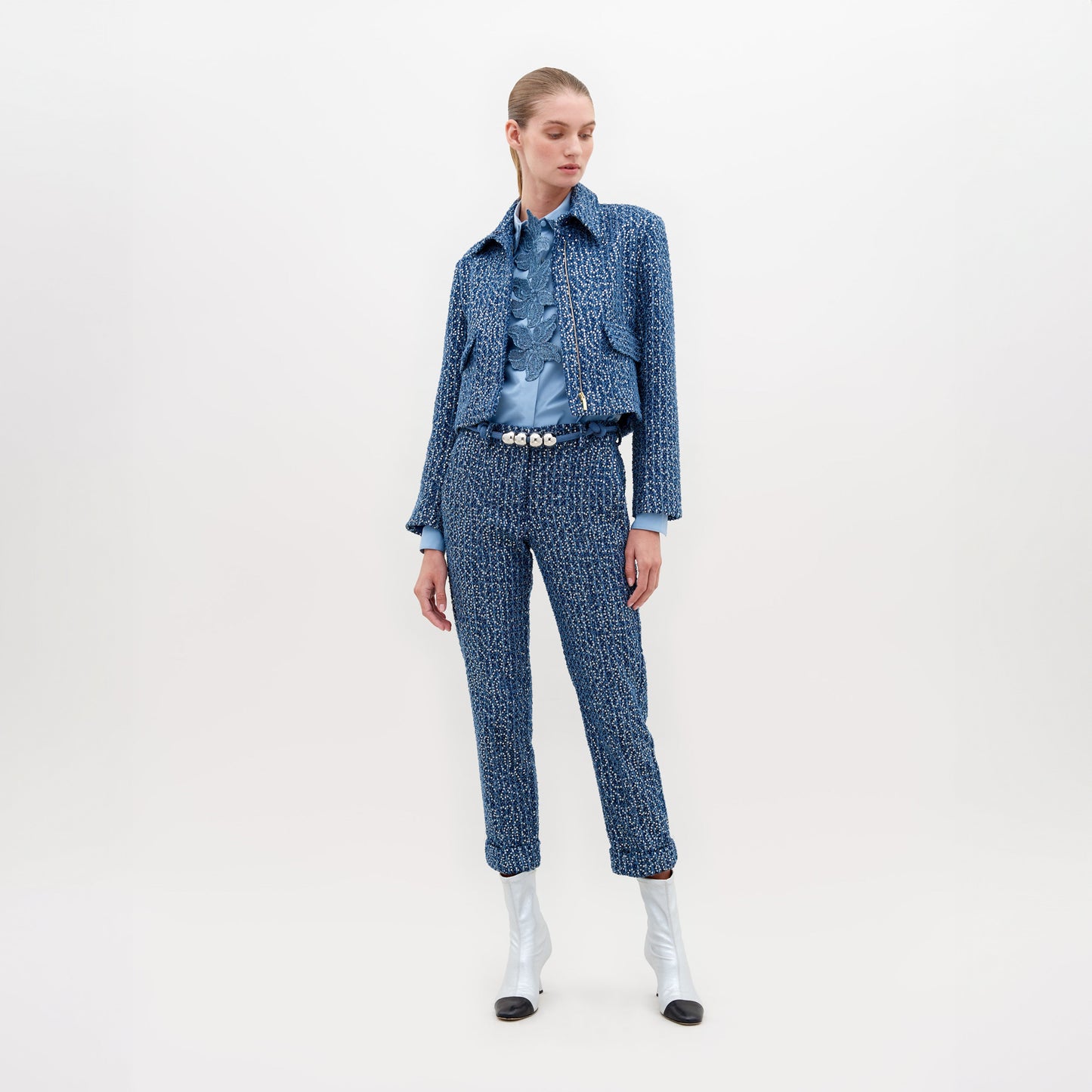 A woman stands against a plain background wearing a white blouse with blue lace detail, Garmet Pant Navy Sequins, a pearl belt, and white ankle boots with black tips. This stylish ensemble is available for pre-order now and will ship by November 15th, 2024.