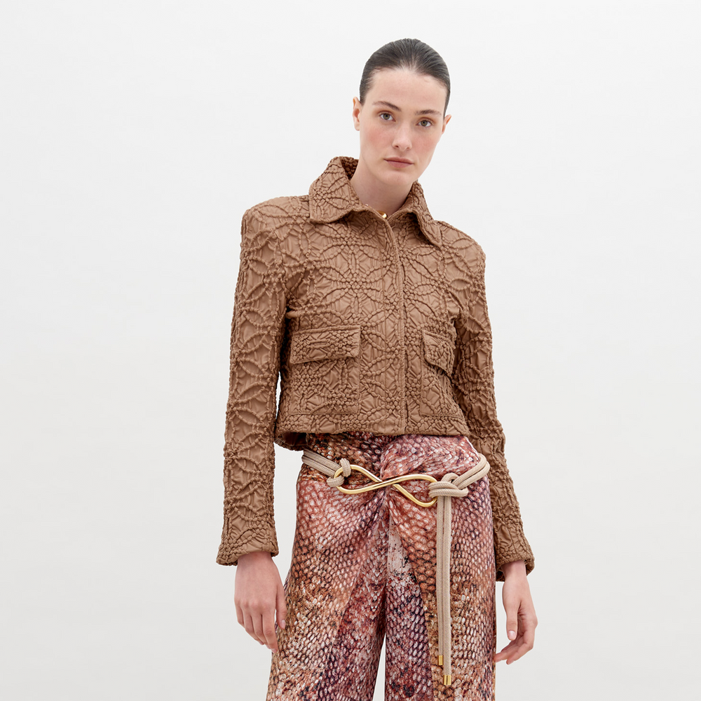 A woman stands against a plain background, wearing the Perla Jacket Brown and patterned high-waist pants. A decorative knot belt accentuates her waist. This stylish ensemble is available now, ensuring you can enhance your wardrobe immediately.