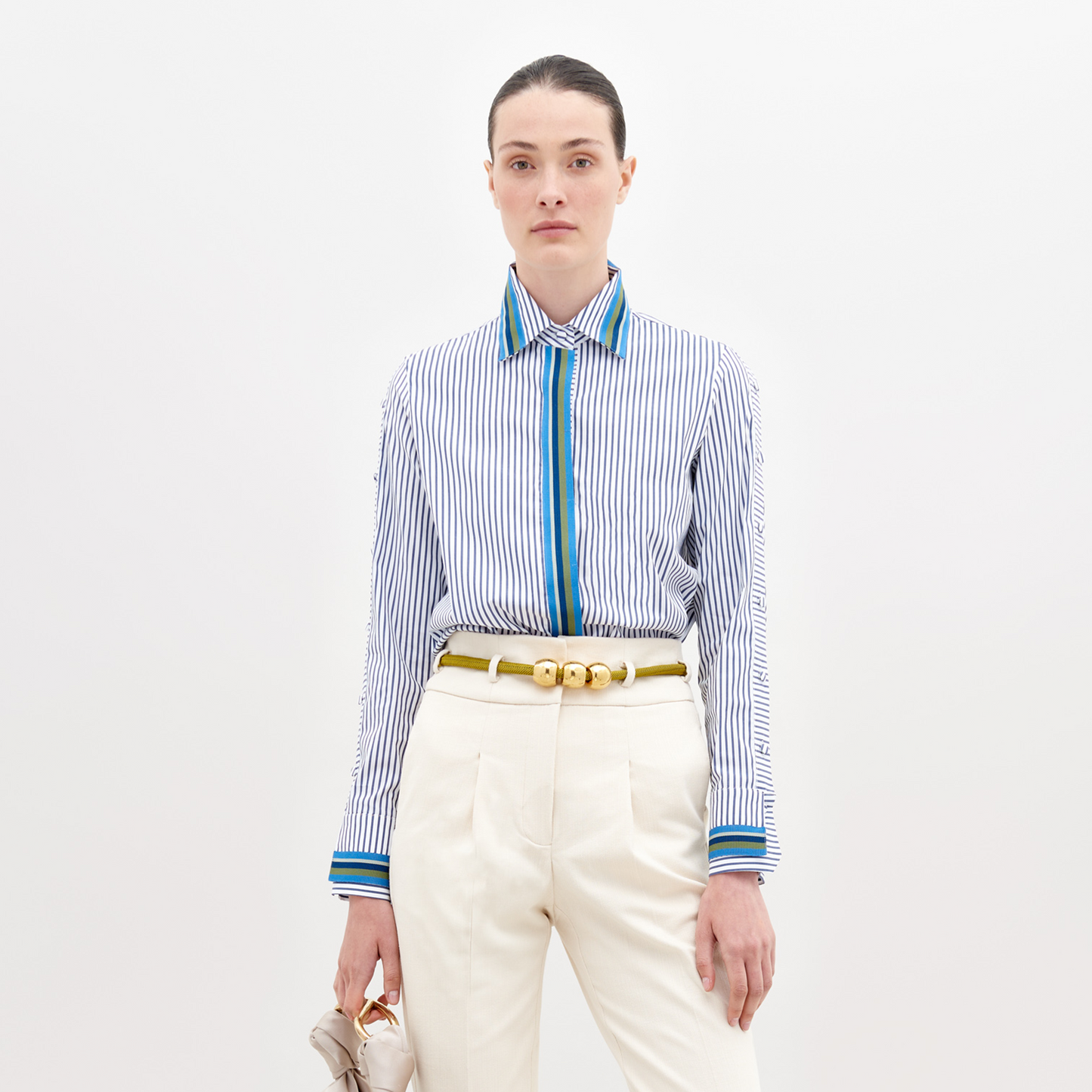 A person wearing a Premium Shirt White Blue Stripes with a blue accent, white pants, and a yellow belt holds a small bag in their hand. The background is plain white. This stylish ensemble will be available for pre-order starting November 15th, 2024.