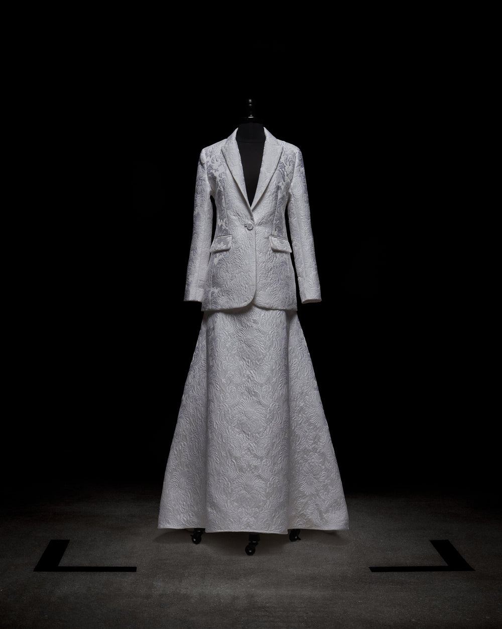 Wearing an Imola Skirt, a person in a white textured suit holds a bouquet of light-colored flowers against a plain background.