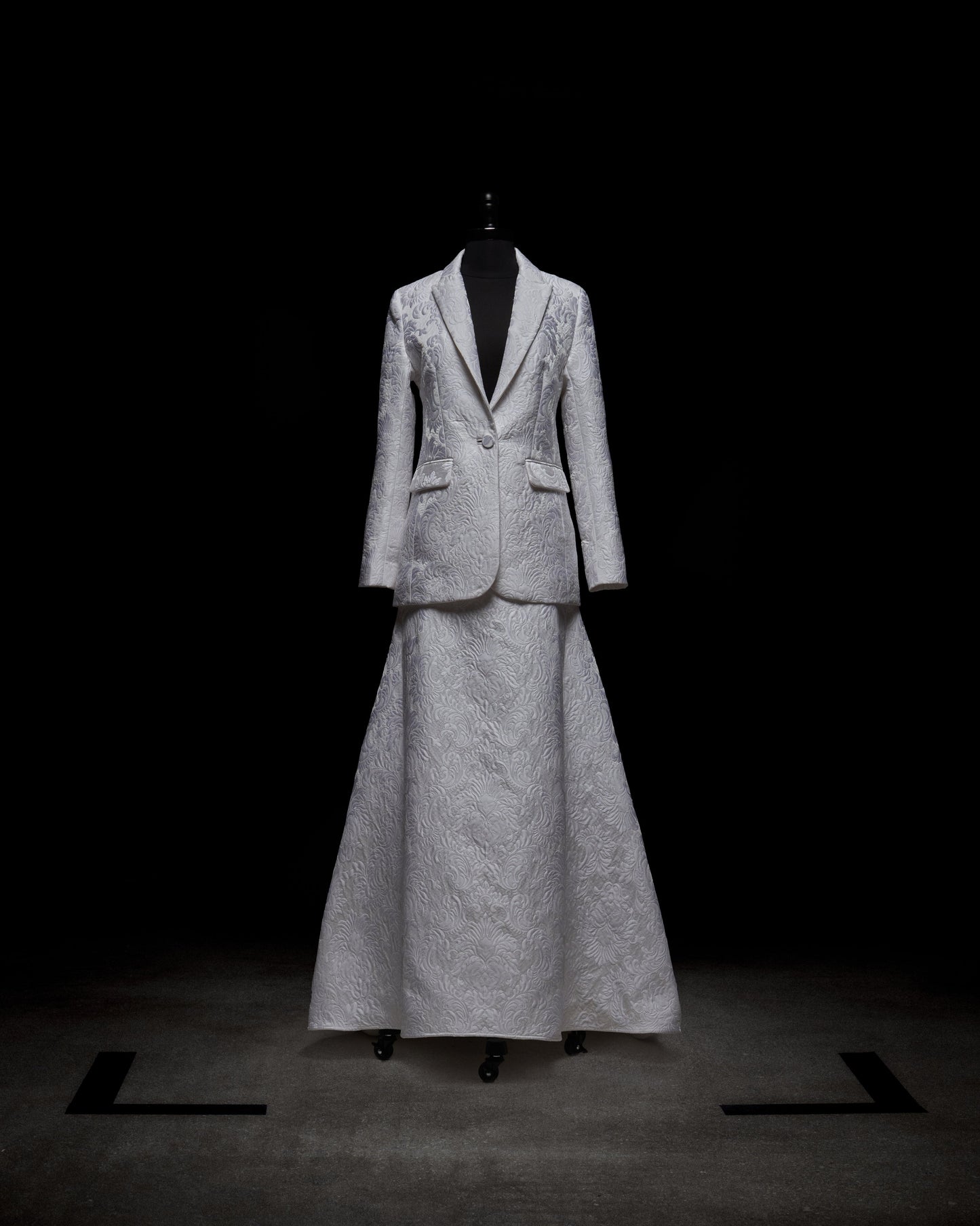 Wearing an Imola Skirt, a person in a white textured suit holds a bouquet of light-colored flowers against a plain background.