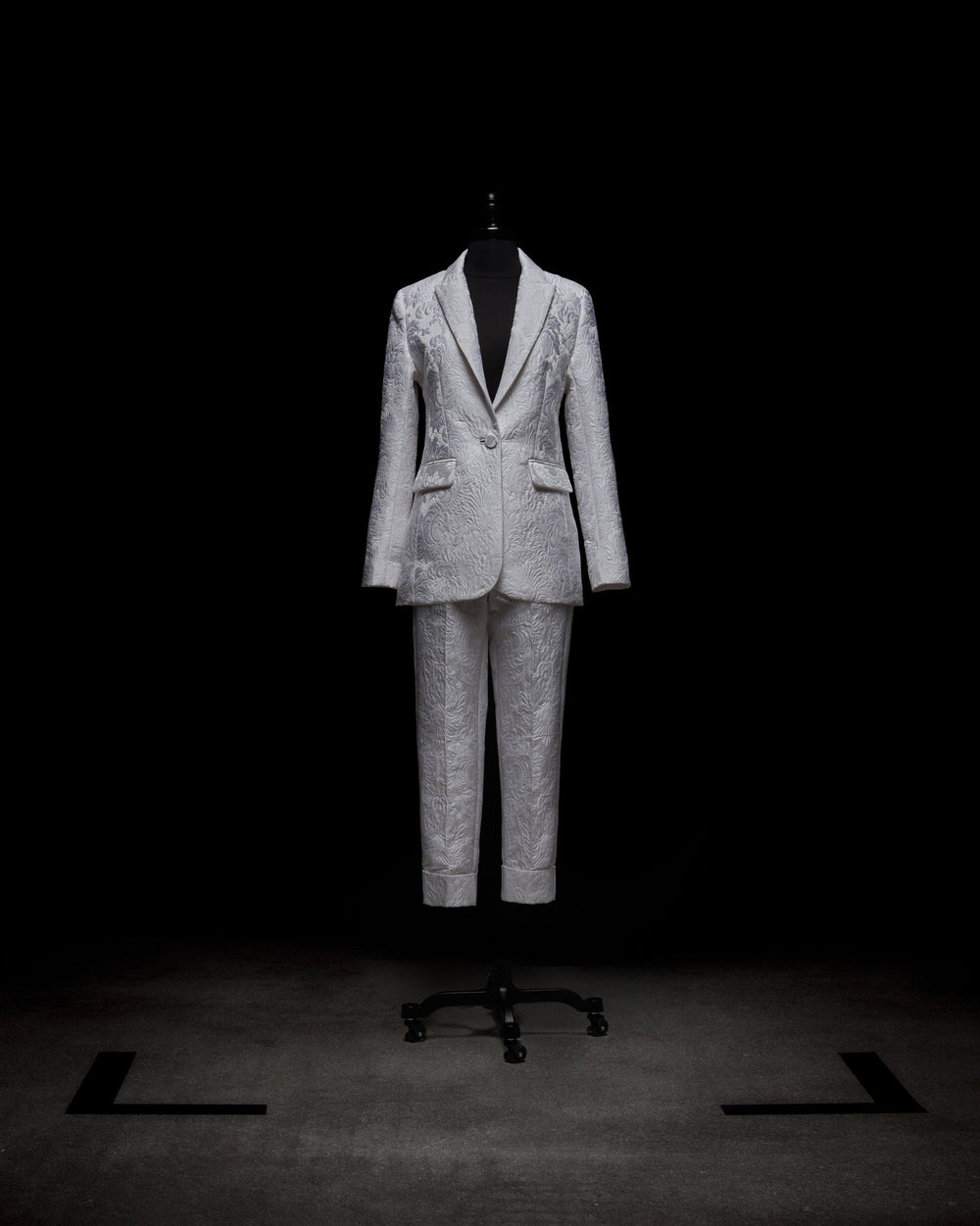 A person in a white Moad Pant suit and boots holds a textured handbag, standing against a plain white background.