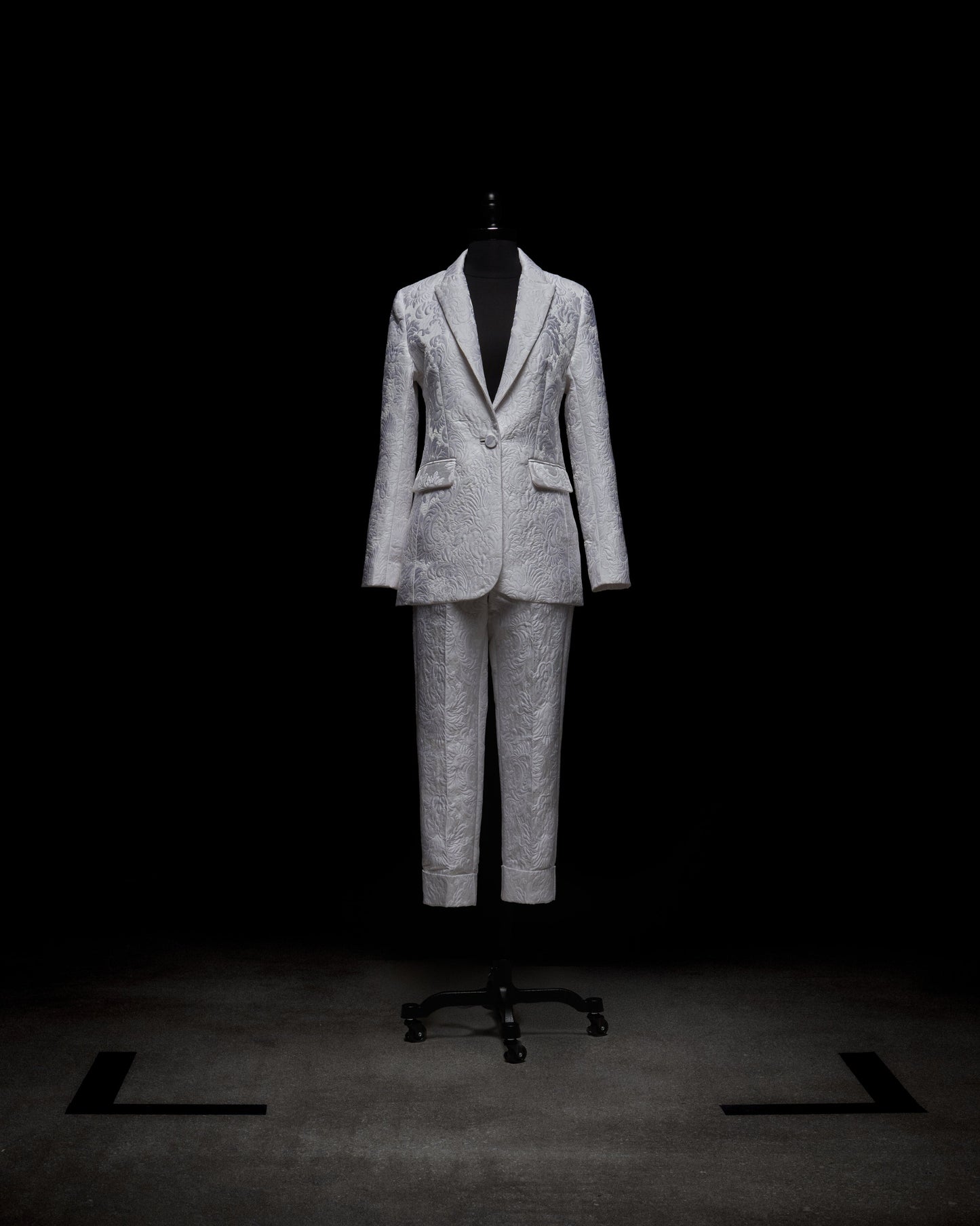 A person in a white Moad Pant suit and boots holds a textured handbag, standing against a plain white background.