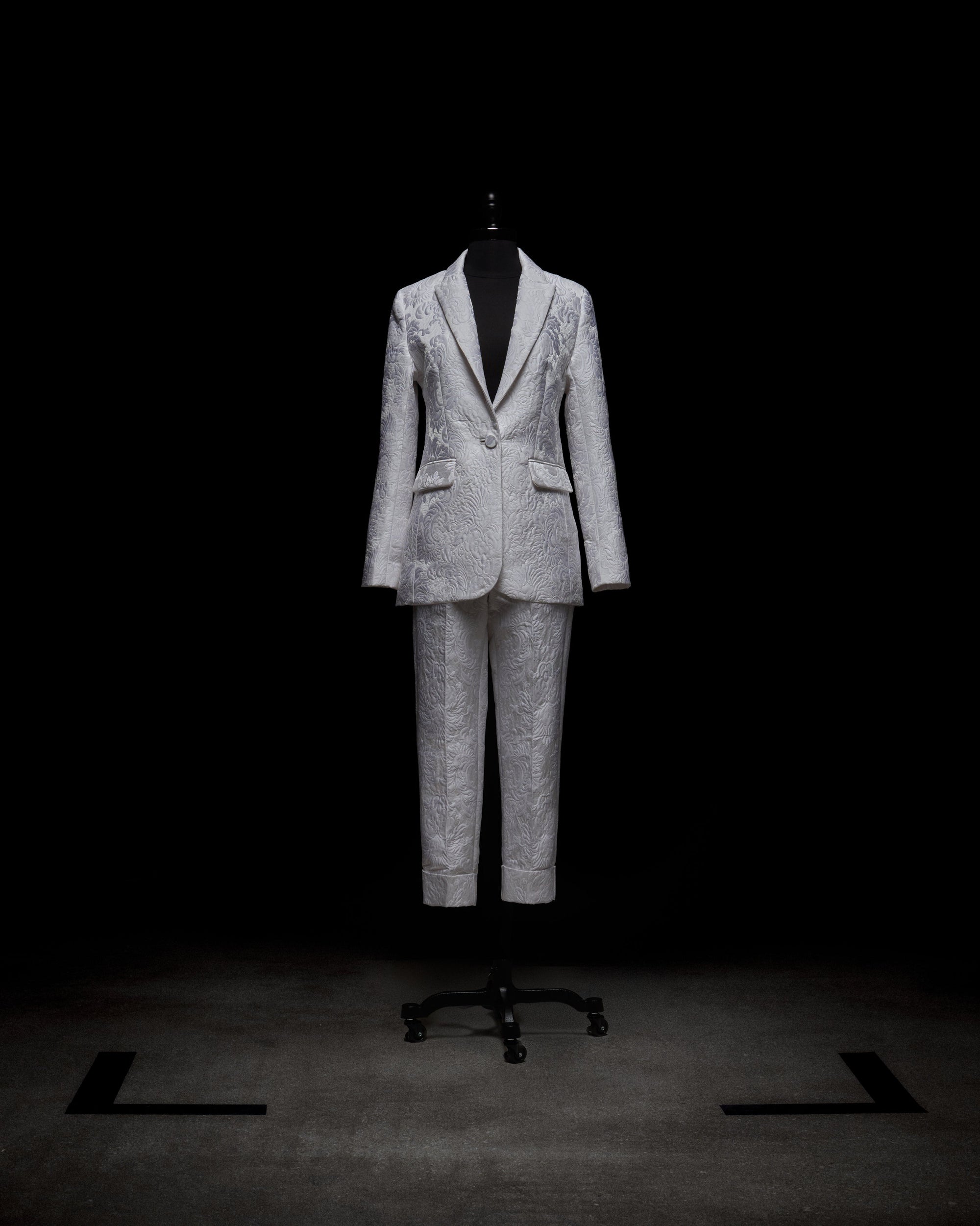 A person in a white Moad Pant suit and boots holds a textured handbag, standing against a plain white background.