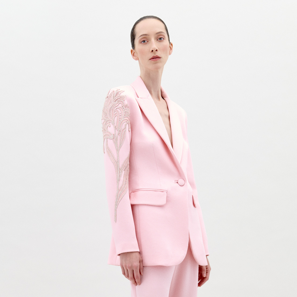 Profile view of a person with long hair tied back, wearing the Rebeca Jacket Pink, a light pink blazer adorned with intricate embroidery on the shoulder; pre-order now for delivery by February 1st, 2025.
