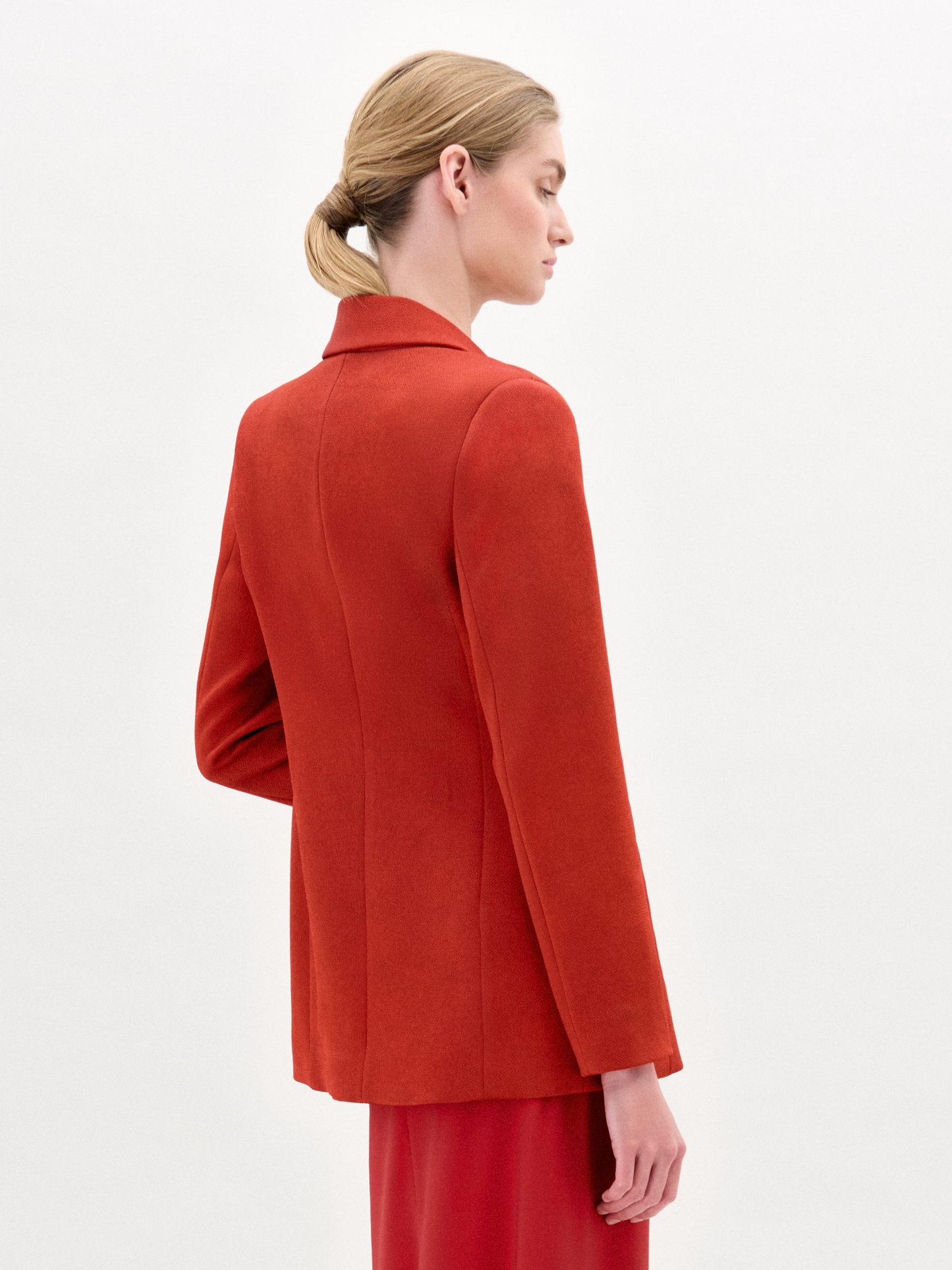 A person wearing the Rebeca Jacket, a fitted red blazer made from Italian fabric, layered over a patterned red and beige dress, stands against a plain light background.