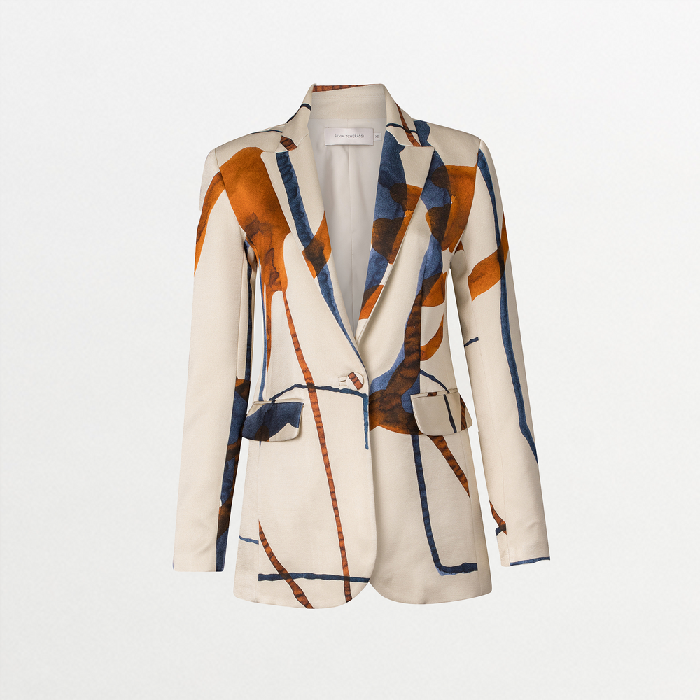 A woman with tied back hair stands against a blue background, wearing the Rebeca Jacket in Cobalt Copper Abstract with a matching dress, and large, abstract earrings, ready for her feature on September 2nd.