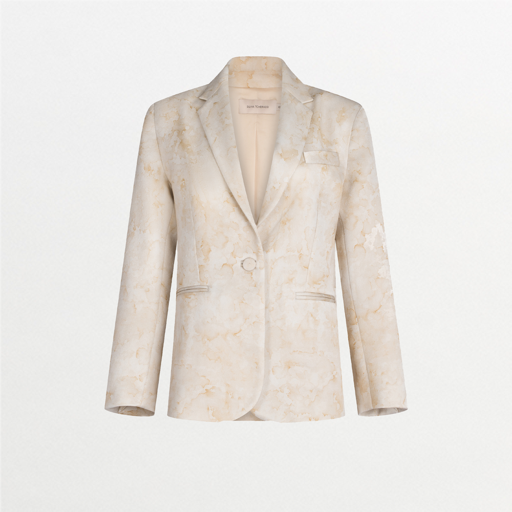 A person stands against a plain blue background wearing the Rebeca Jacket Cream Abstract Print draped over their shoulders, complemented by a white blouse with a ruffled collar, matching pants, and a gold belt with an "S" charm. The ensemble exudes timeless elegance, perfect for events like August 19th or September 2nd.