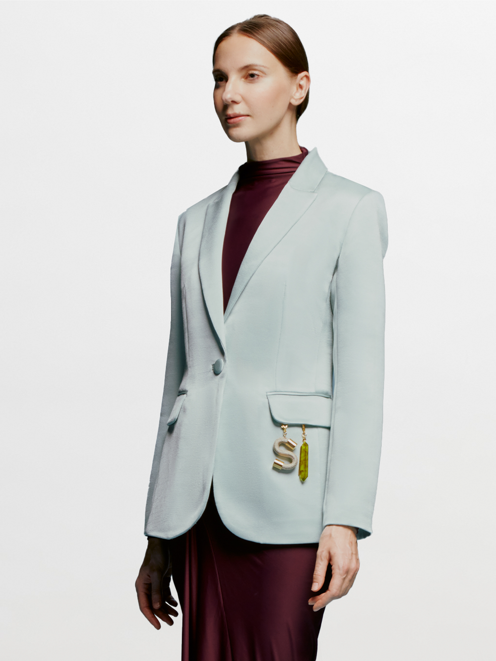A woman in a turquoise Rebeca Jacket over a burgundy top stands against a light blue background. She has straight, pulled-back hair and an "S" charm with an attached trinket hanging from her jacket pocket, ready to make a statement on September 2nd.