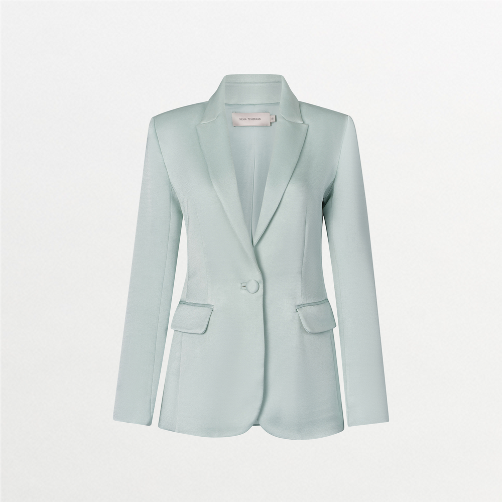 A woman in a turquoise Rebeca Jacket over a burgundy top stands against a light blue background. She has straight, pulled-back hair and an "S" charm with an attached trinket hanging from her jacket pocket, ready to make a statement on September 2nd.