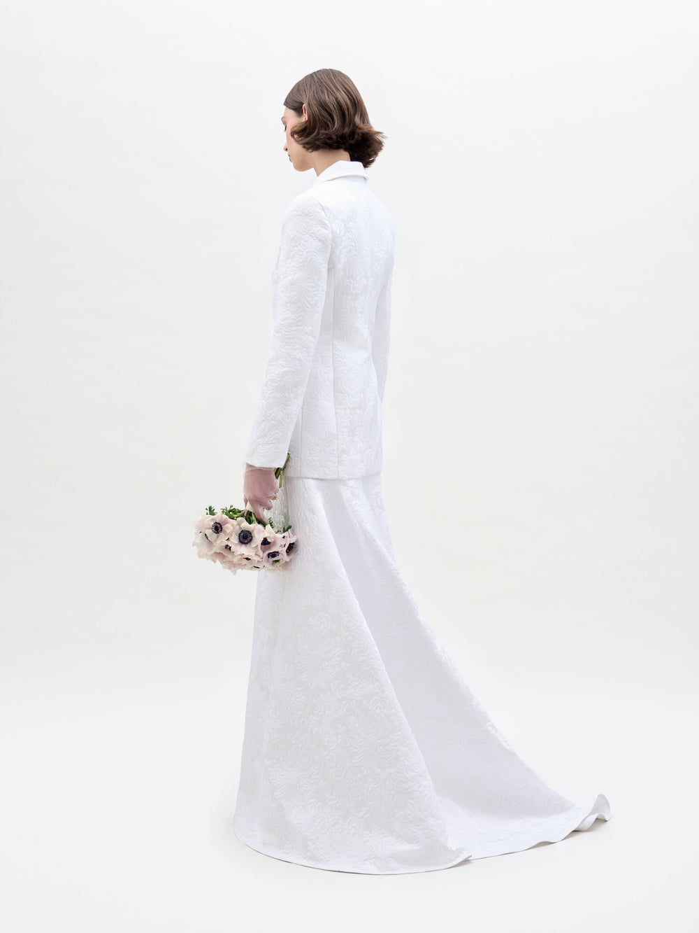 Wearing an Imola Skirt, a person in a white textured suit holds a bouquet of light-colored flowers against a plain background.