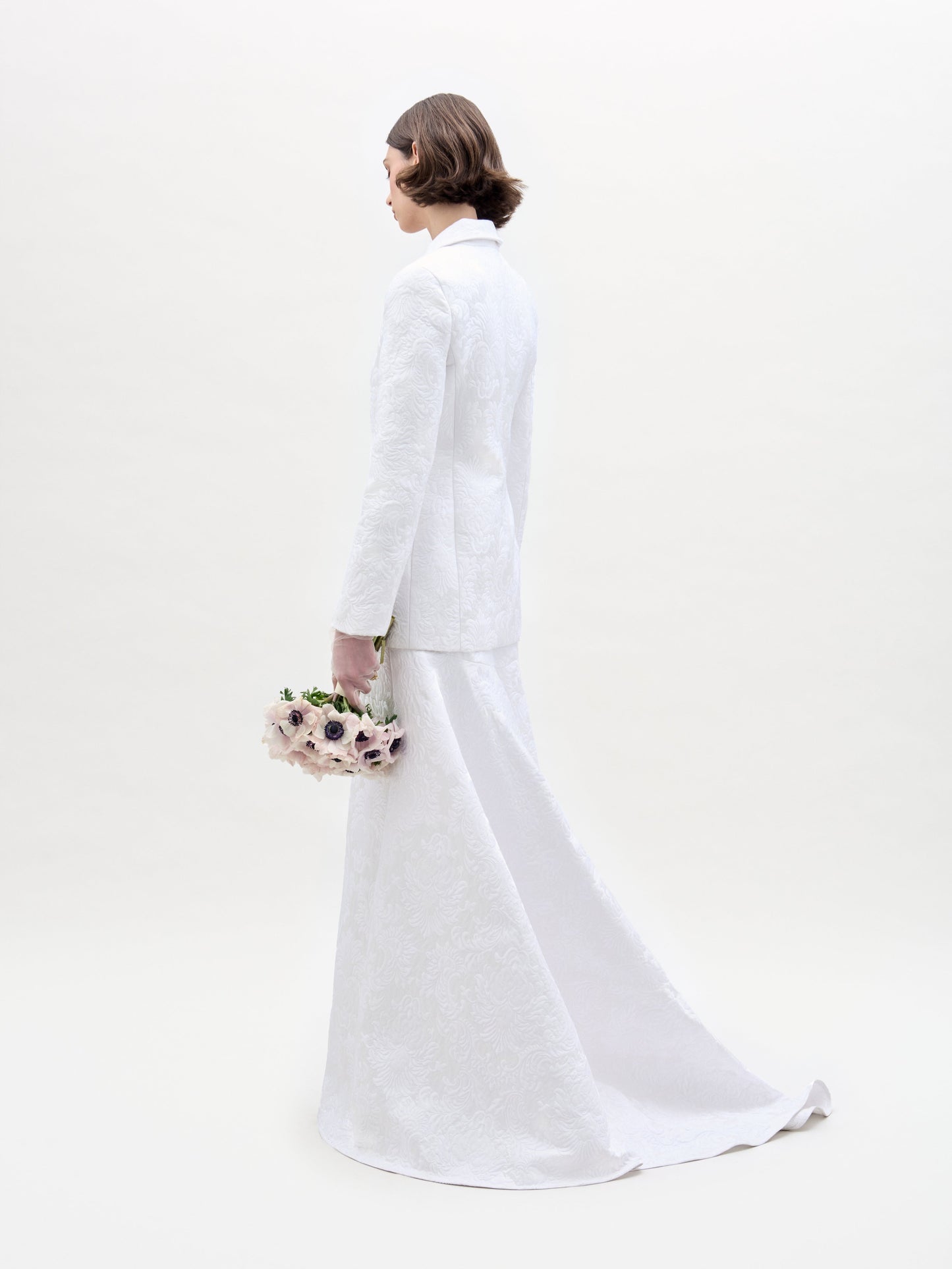 Wearing an Imola Skirt, a person in a white textured suit holds a bouquet of light-colored flowers against a plain background.