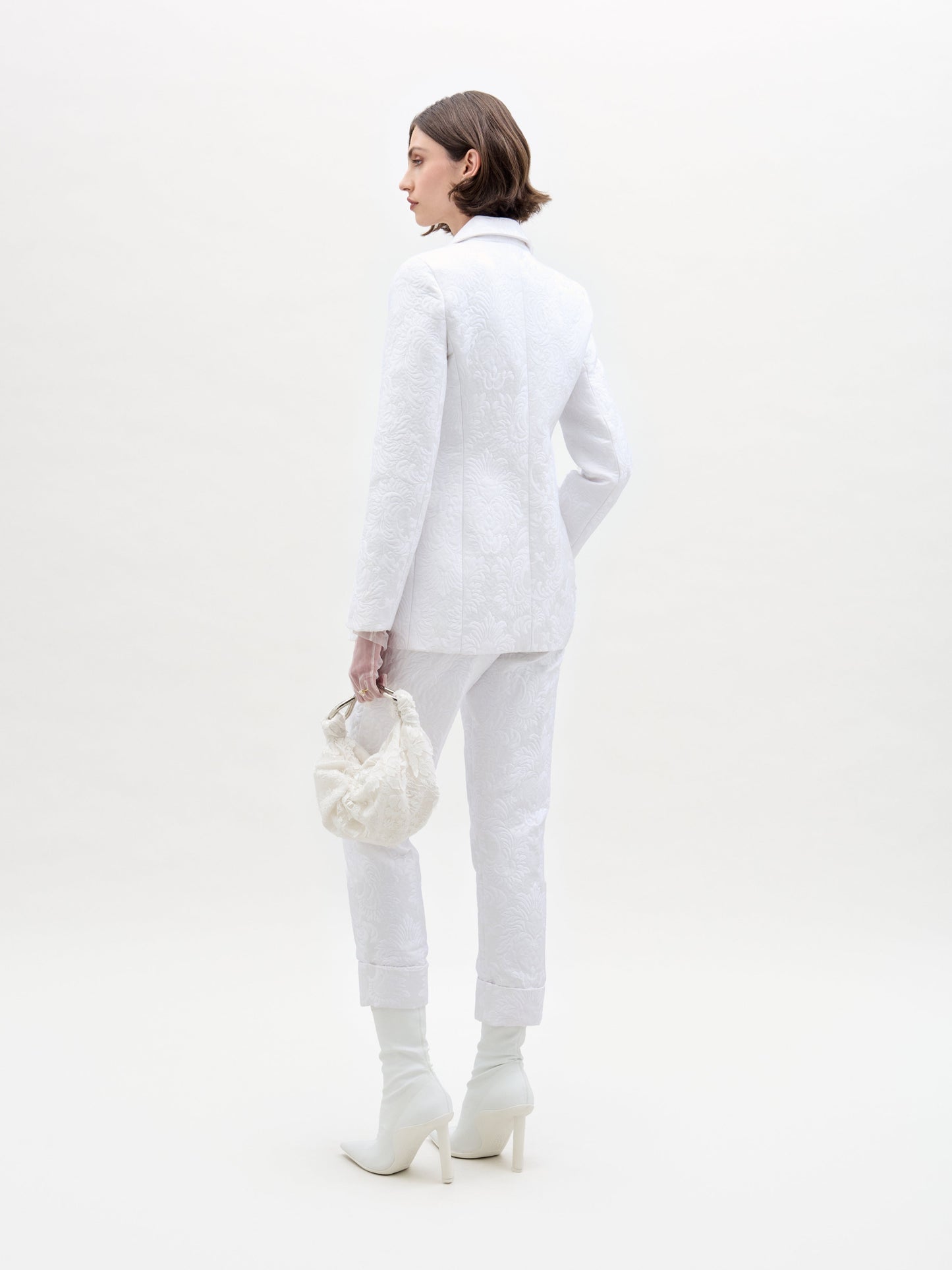 A person in a white Moad Pant suit and boots holds a textured handbag, standing against a plain white background.
