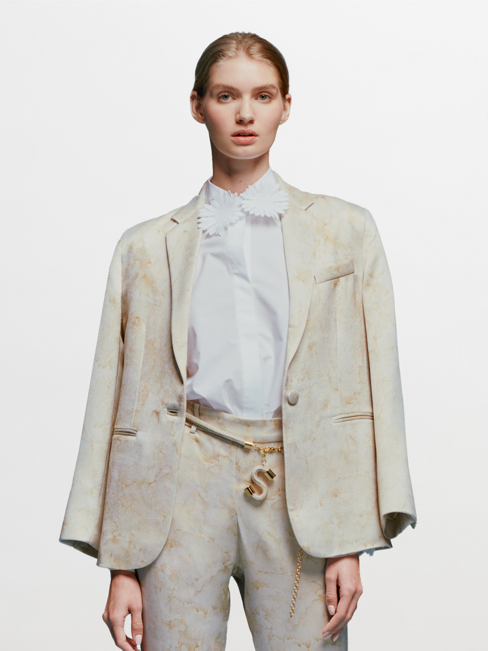 A person stands against a plain blue background wearing the Rebeca Jacket Cream Abstract Print draped over their shoulders, complemented by a white blouse with a ruffled collar, matching pants, and a gold belt with an "S" charm. The ensemble exudes timeless elegance, perfect for events like August 19th or September 2nd.