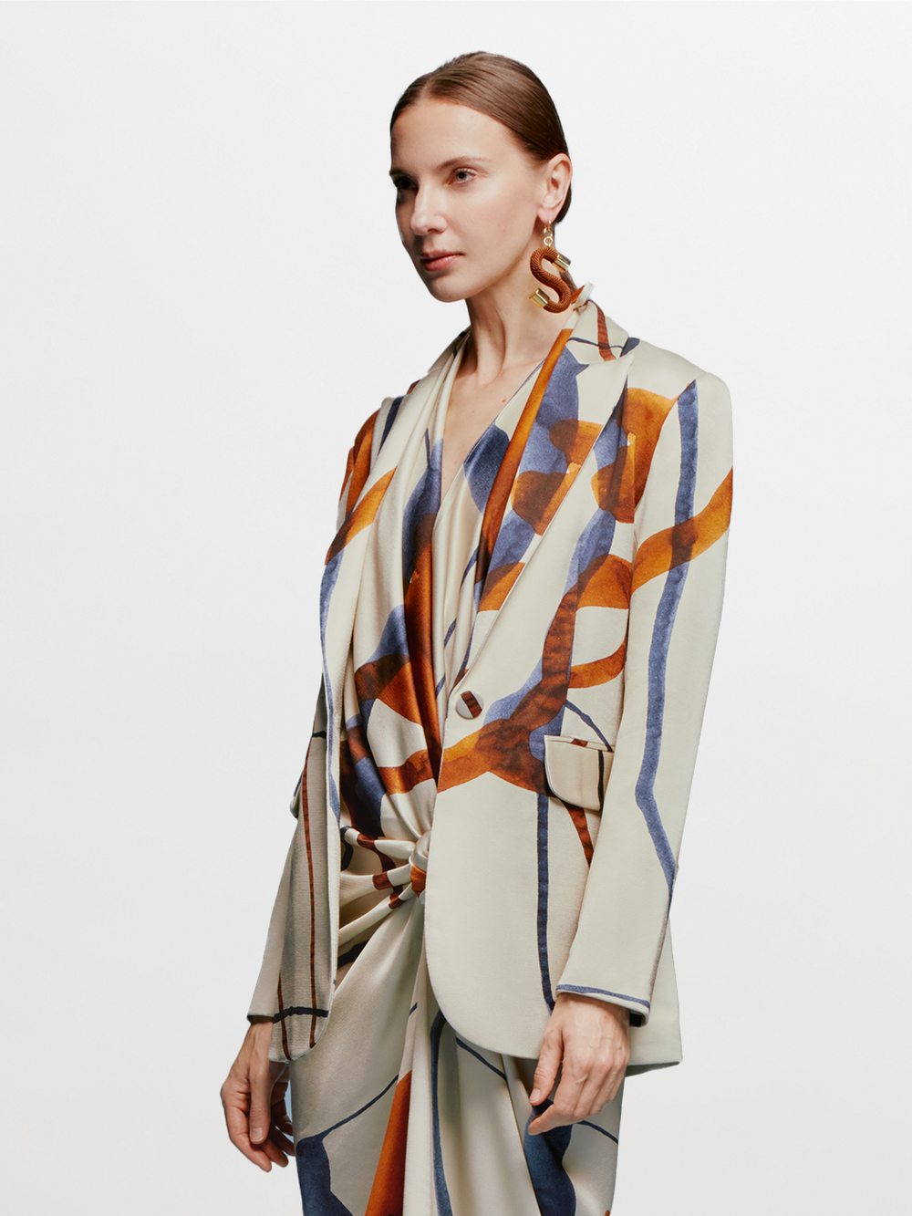 A woman with tied back hair stands against a blue background, wearing the Rebeca Jacket in Cobalt Copper Abstract with a matching dress, and large, abstract earrings, ready for her feature on September 2nd.