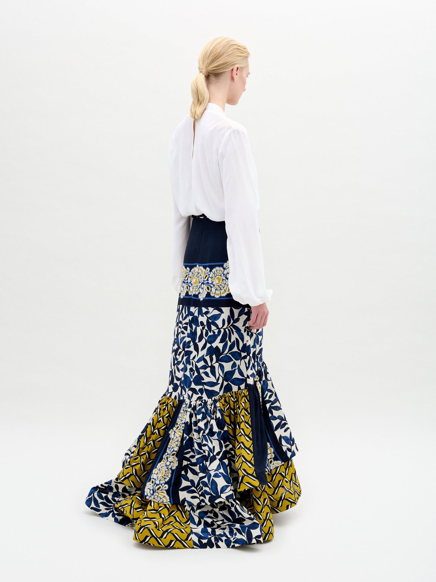 Model wearing the Rocio Blouse—a white, collarless V-neck top—paired with a floral belt featuring a gold buckle and a navy Italian fabric skirt with yellow and white floral prints, standing against a plain background.