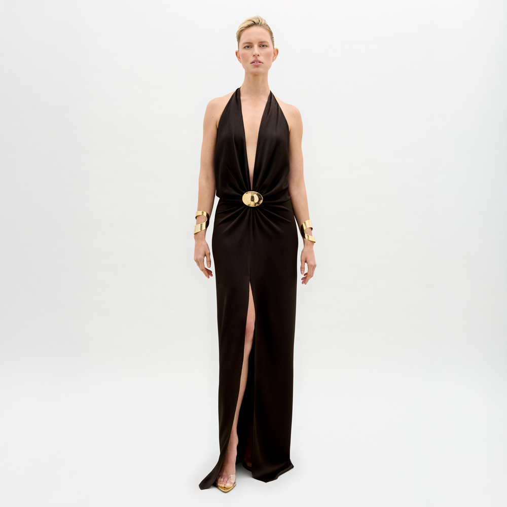 A person stands against a white background in the Rowan Dress, featuring a sleeveless deep V-neck black hammered satin maxi silhouette with a central gold embellishment, paired with gold bracelets, short hair, and gold high-heeled shoes.
