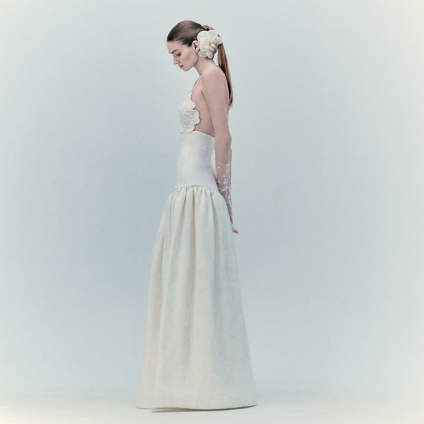 A woman in the Ruth Dress Pearl, a long white maxi, stands in profile against a plain light background with her hair tied back and adorned by a large white flower.