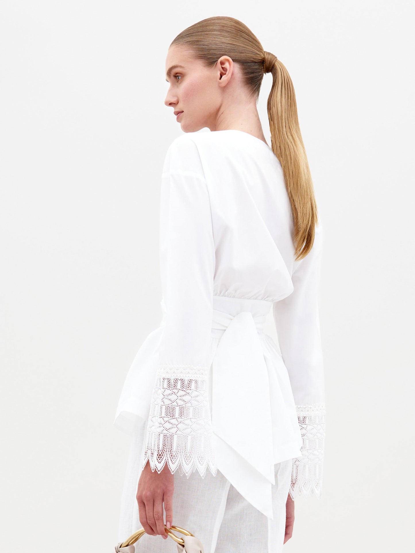 Person wearing the Saanvi Blouse White with lace detail and coordinating white pants, holding a small handbag, looking off to the side against a plain background. Don't miss your chance to pre-order this stunning outfit by the ship-by date of February 15th, 2025.