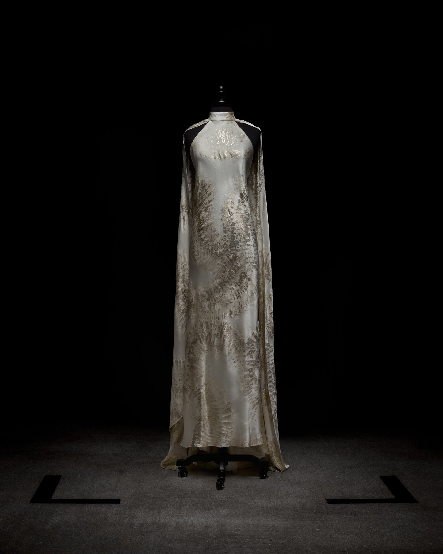 A woman poses in the Sabina Dress, a white and beige flowing gown featuring a high neckline and cape-like sleeves, set against a plain backdrop.