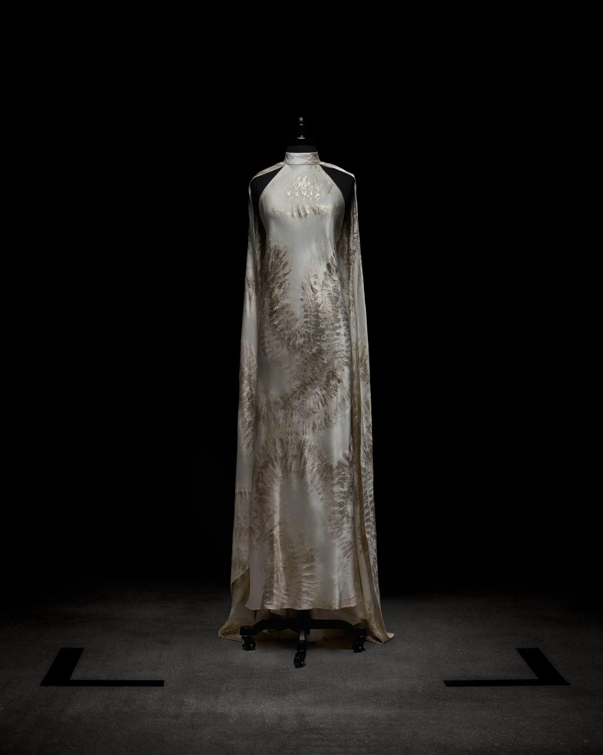 A woman poses in the Sabina Dress, a white and beige flowing gown featuring a high neckline and cape-like sleeves, set against a plain backdrop.