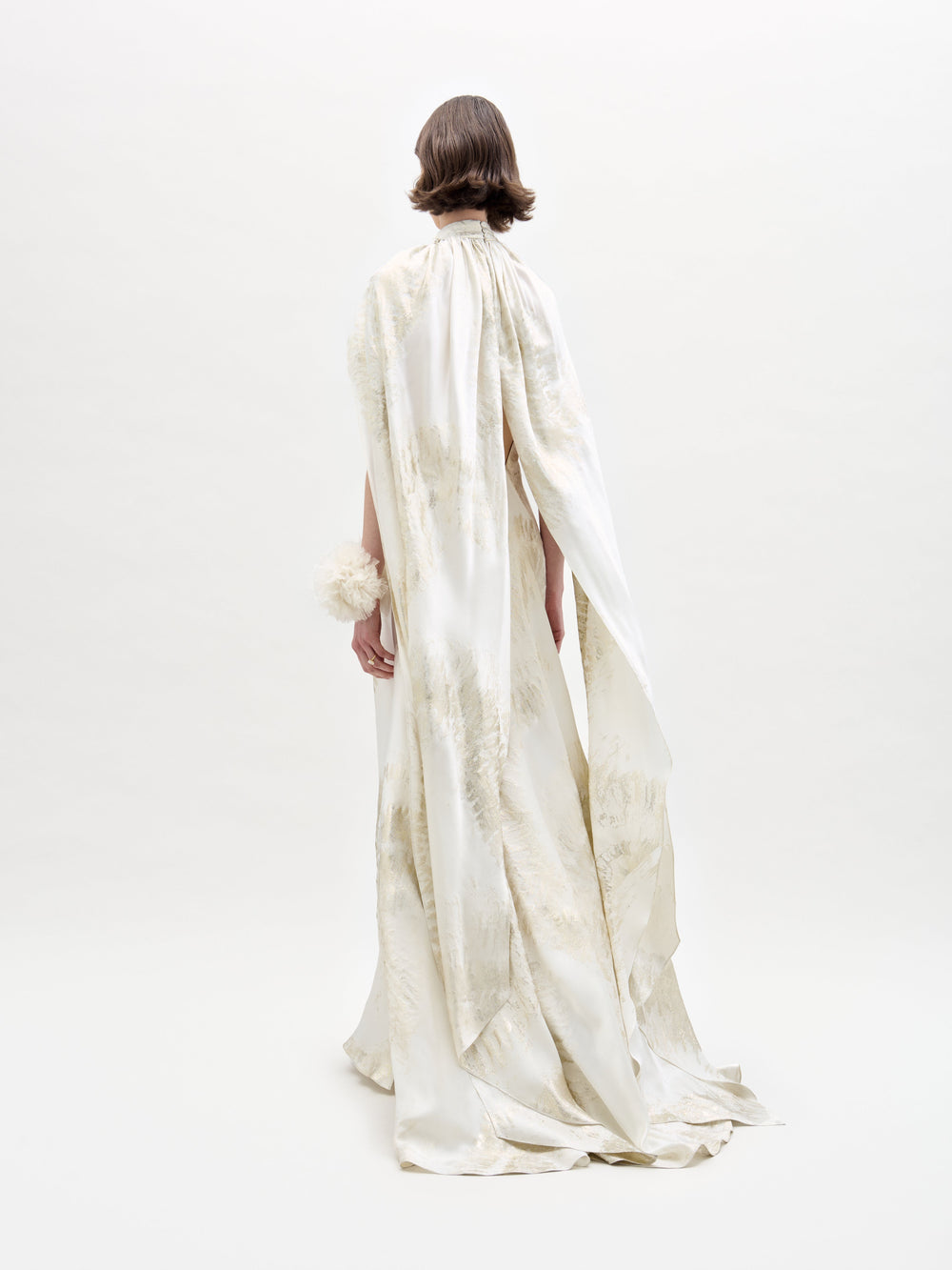 A woman poses in the Sabina Dress, a white and beige flowing gown featuring a high neckline and cape-like sleeves, set against a plain backdrop.