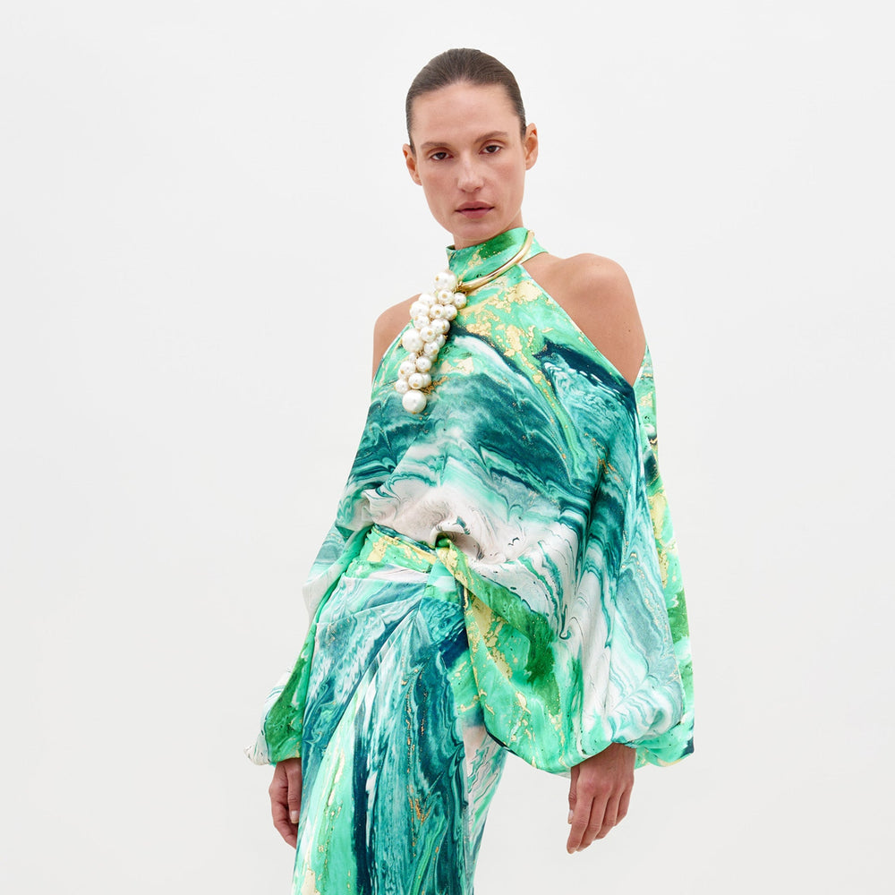 A person wearing the Safira Blouse Green Marble, a sophisticated green and white marble-patterned outfit with shoulder cutouts, accessorized with a large pearl necklace, stands against a plain white background. Note: Available for pre-order, ships by February 15th.