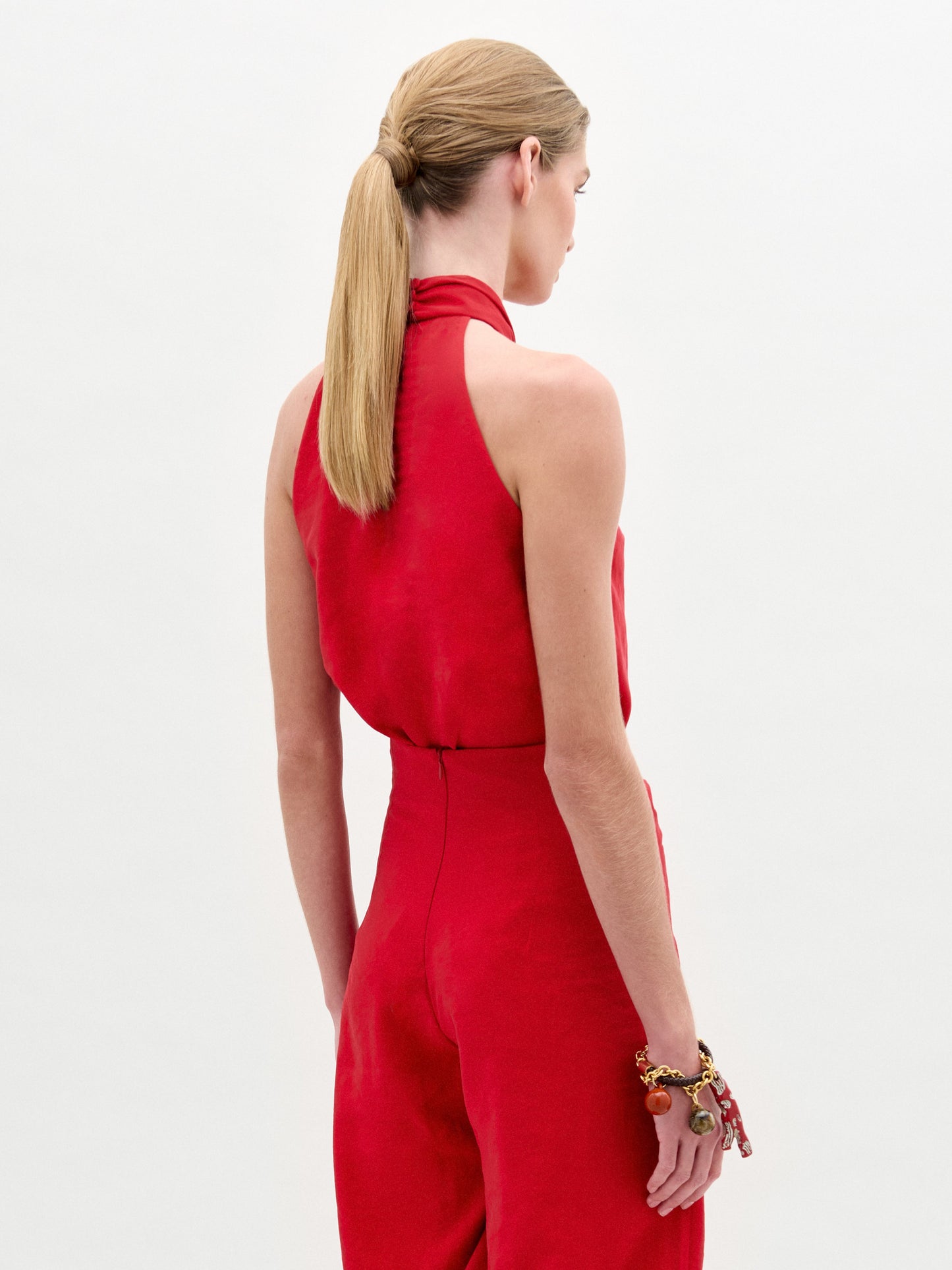 Woman wearing the Shela Blouse, a sleeveless red halter-neck top made from Italian fabric with a knotted front detail, standing against a plain light background.