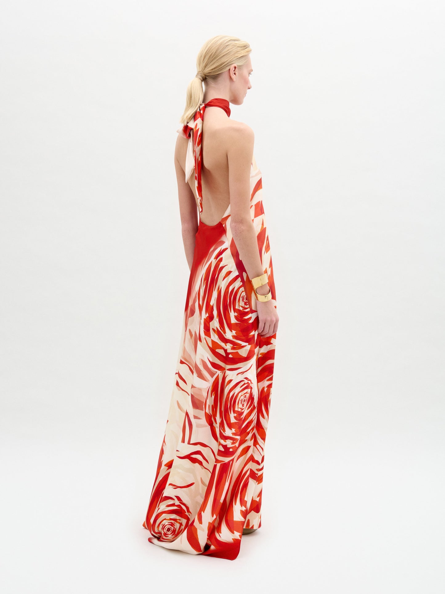 A person stands against a white background wearing the Sherry Dress—a red and white silk maxi with a bold swirling rose pattern, halter neckline, draped column fit—styled with gold accessories.