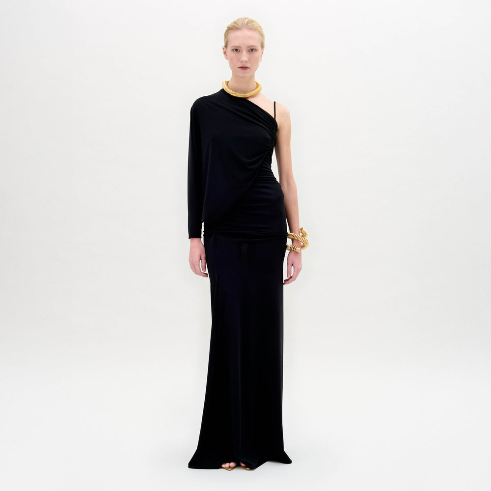 Against a plain background, a person showcases the Shiri Dress, a long black asymmetrical column silhouette crafted from Italian fabric, paired with gold accessories.