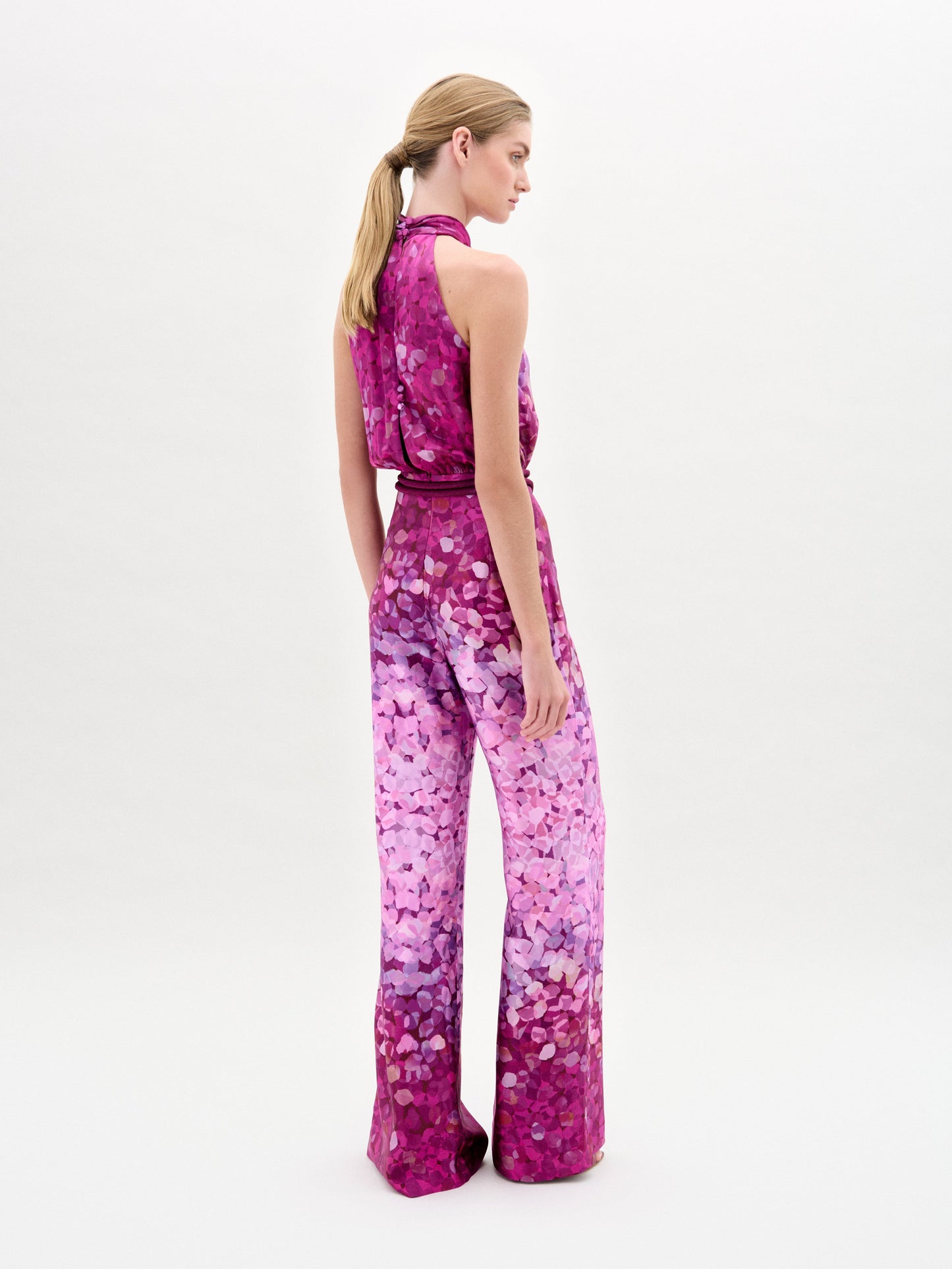 A woman stands against a plain white background wearing the Siv Jumpsuit, a sleeveless halter-neck style made from Italian fabric with a pink and purple abstract print and a matching belt.