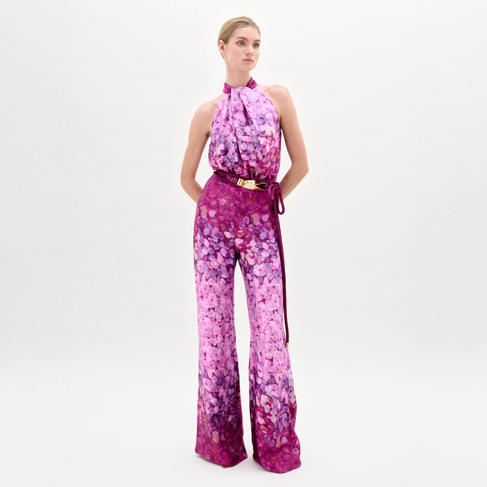 A woman stands against a plain white background wearing the Siv Jumpsuit, a sleeveless halter-neck style made from Italian fabric with a pink and purple abstract print and a matching belt.