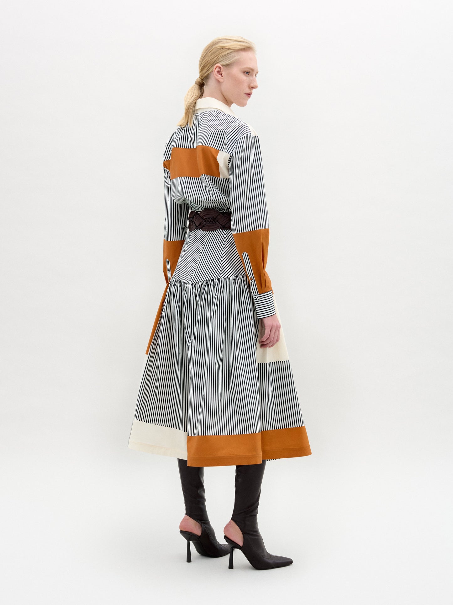 A woman models the Shoreh Skirt, featuring vertical stripes and orange color blocks in Italian fabric. She styles it with a wide gold-buckle belt and knee-high black boots, standing against a plain white background.