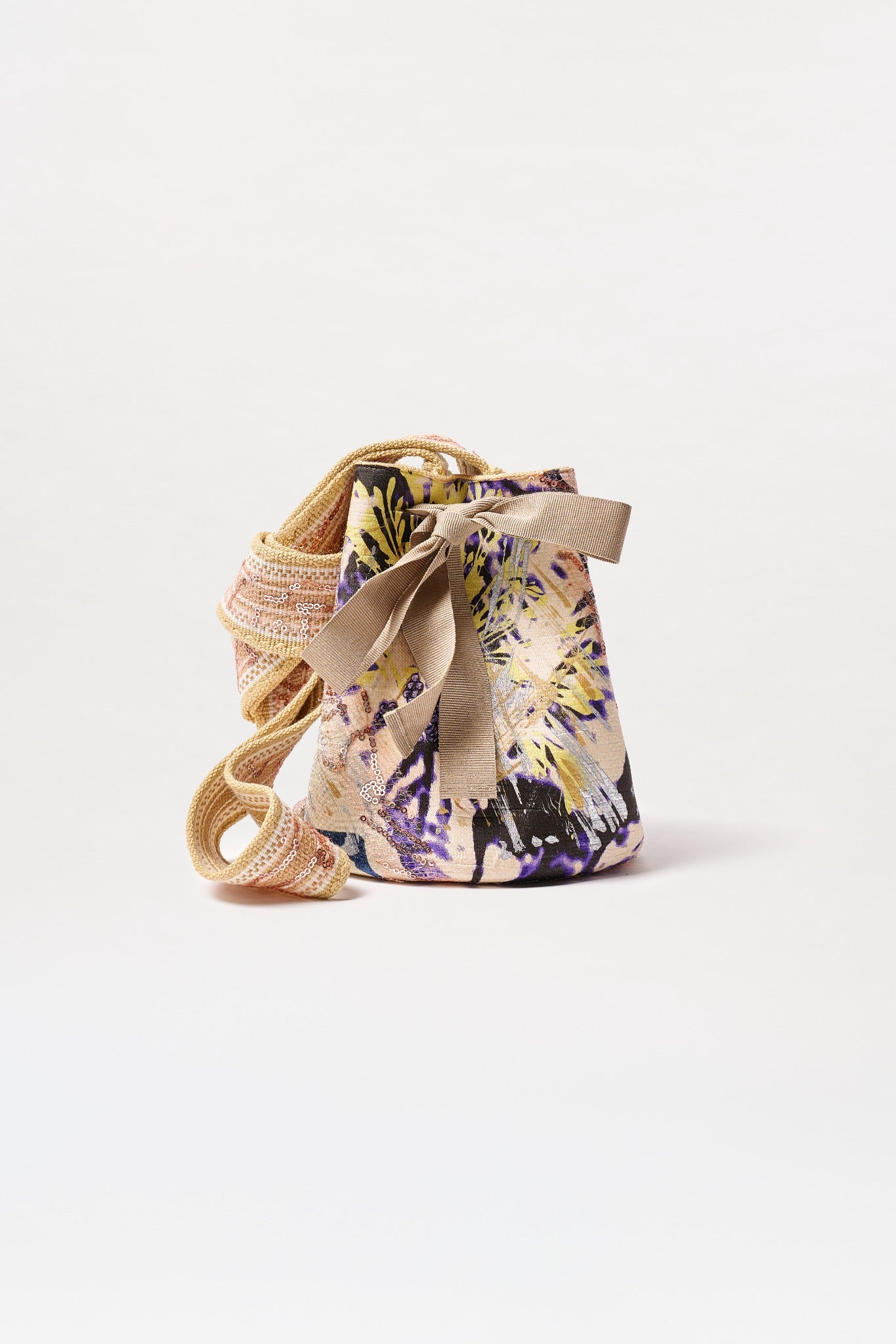 The Mochila Laaput Small is a fabric bucket bag with a colorful abstract floral print, front bow, drawstring closure, and thick textured strap coiled beside it, displayed against a plain white background.