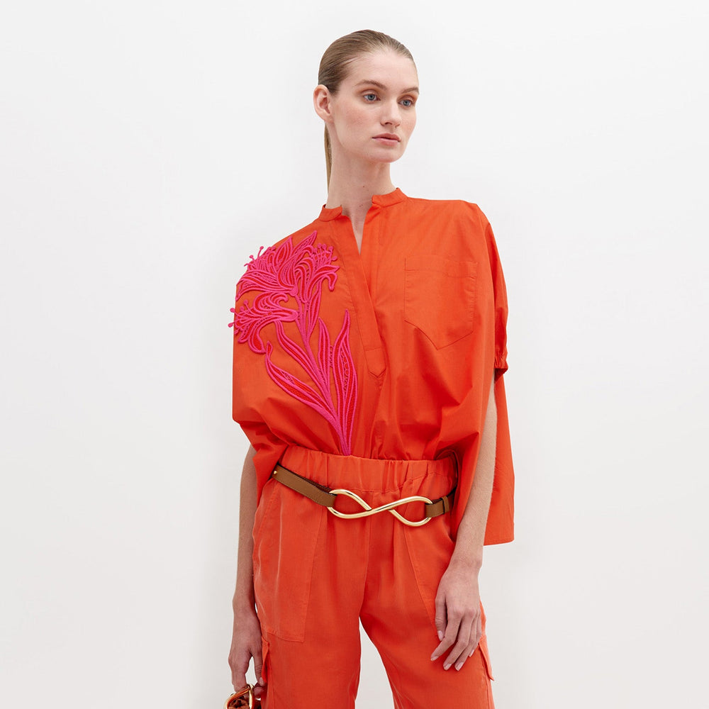 A woman stands against a white background, wearing an orange outfit with a pink floral pattern on the upper left side and a gold belt. The Susanne Blouse Scarlet is available for pre-order until November 15th, 2024.